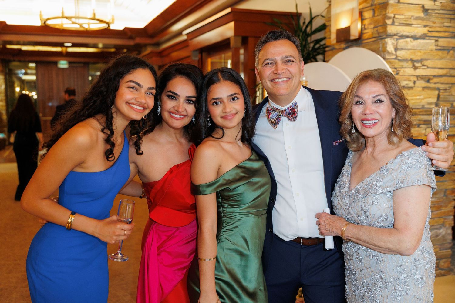 A group in formal attire including blue, red, and green dresses poses together at an elegant venue with stone walls.