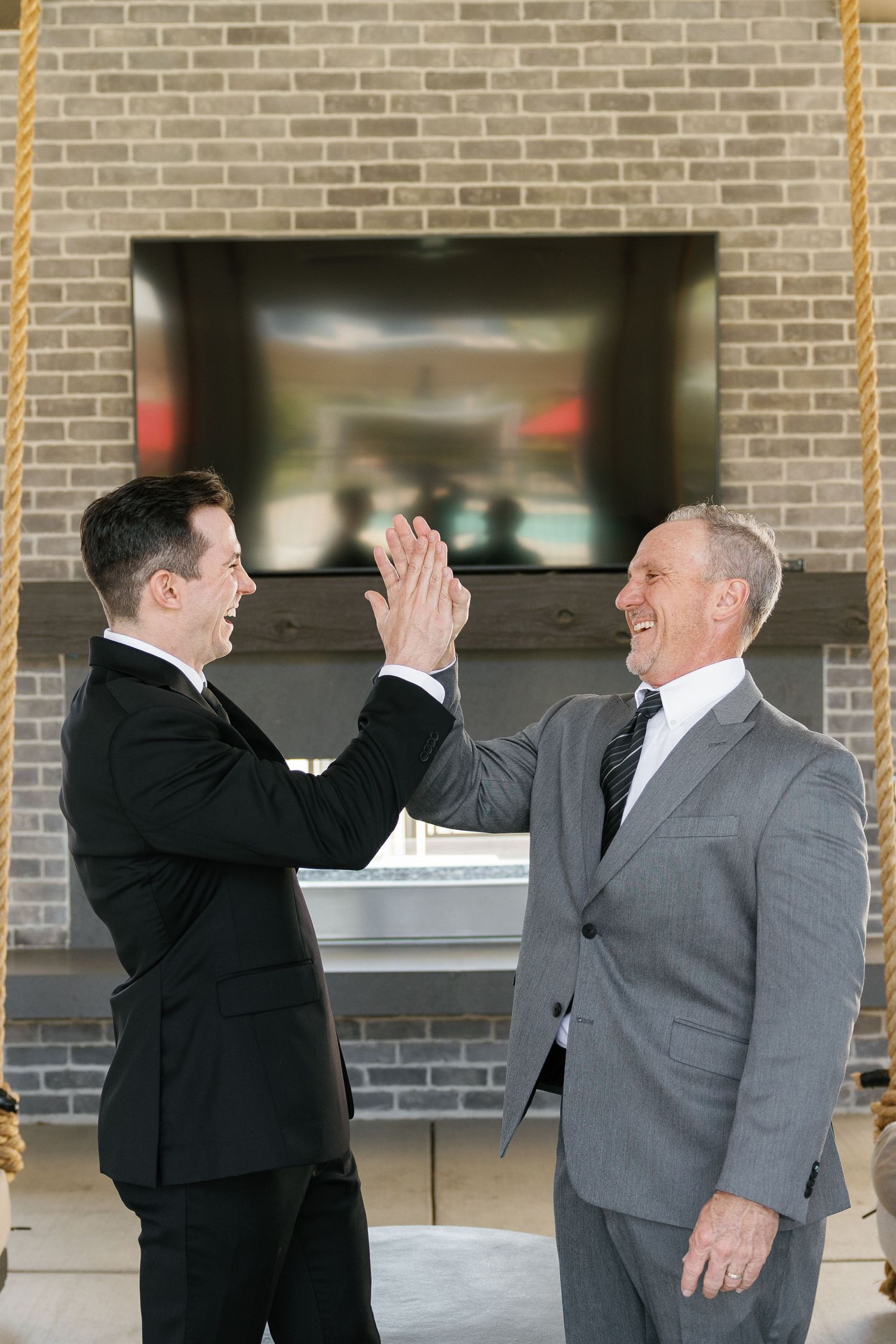 Two businessmen in suits demonstrate a high-five sequence in four progressive frames against a brick wall background.