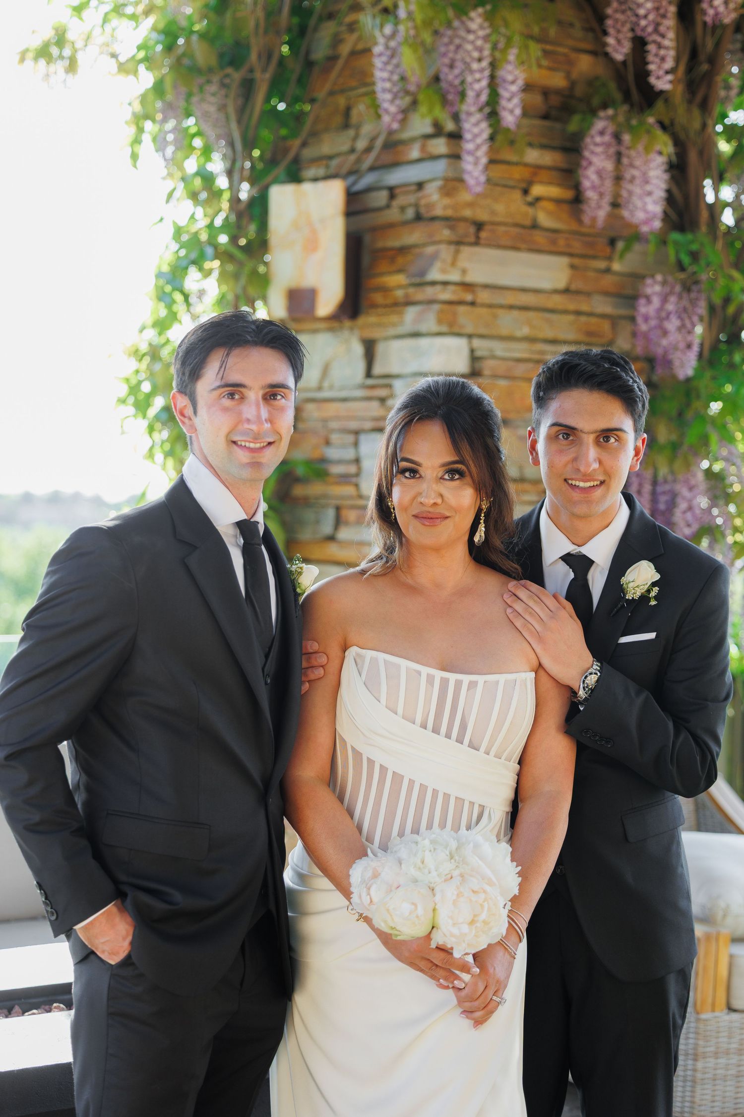 A wedding party poses together under blooming wisteria against a stone wall on a sunny day.