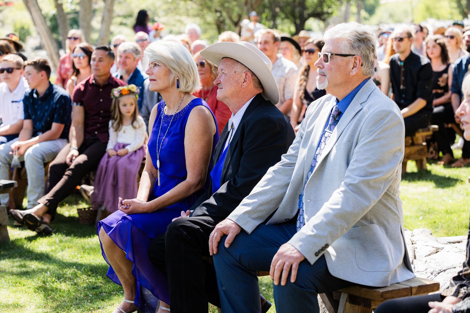 Guests seated in the front row at an outdoor wedding ceremony on a sunny day in a garden setting.
