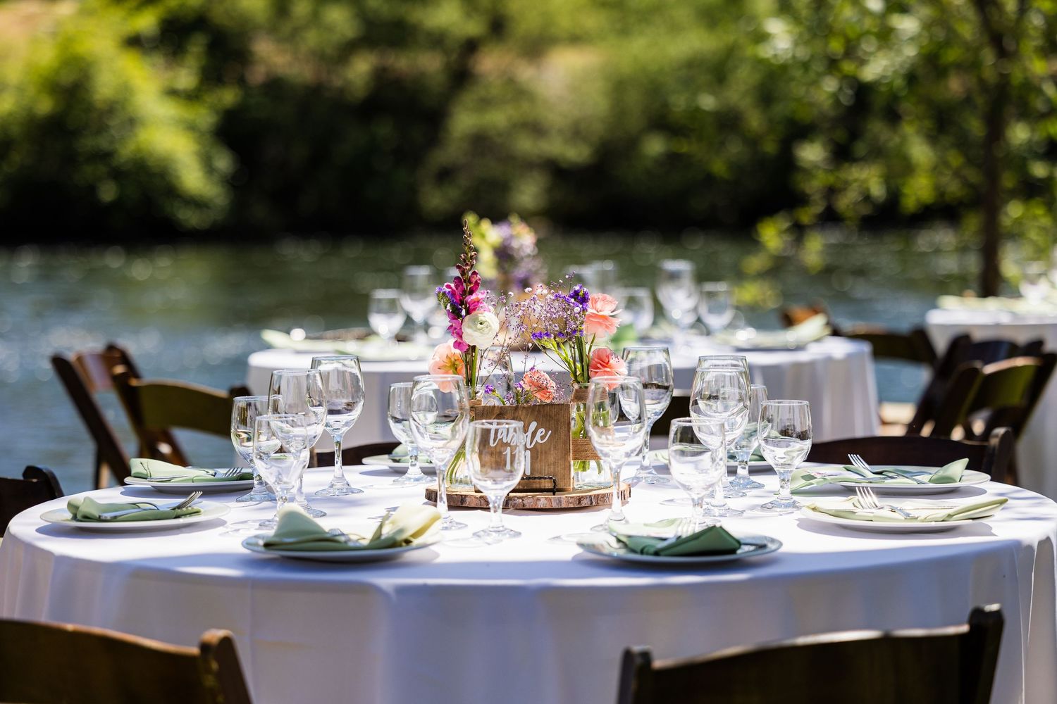 An elegant outdoor table setting with white linens, glassware and floral centerpiece beside a lake in natural lighting.