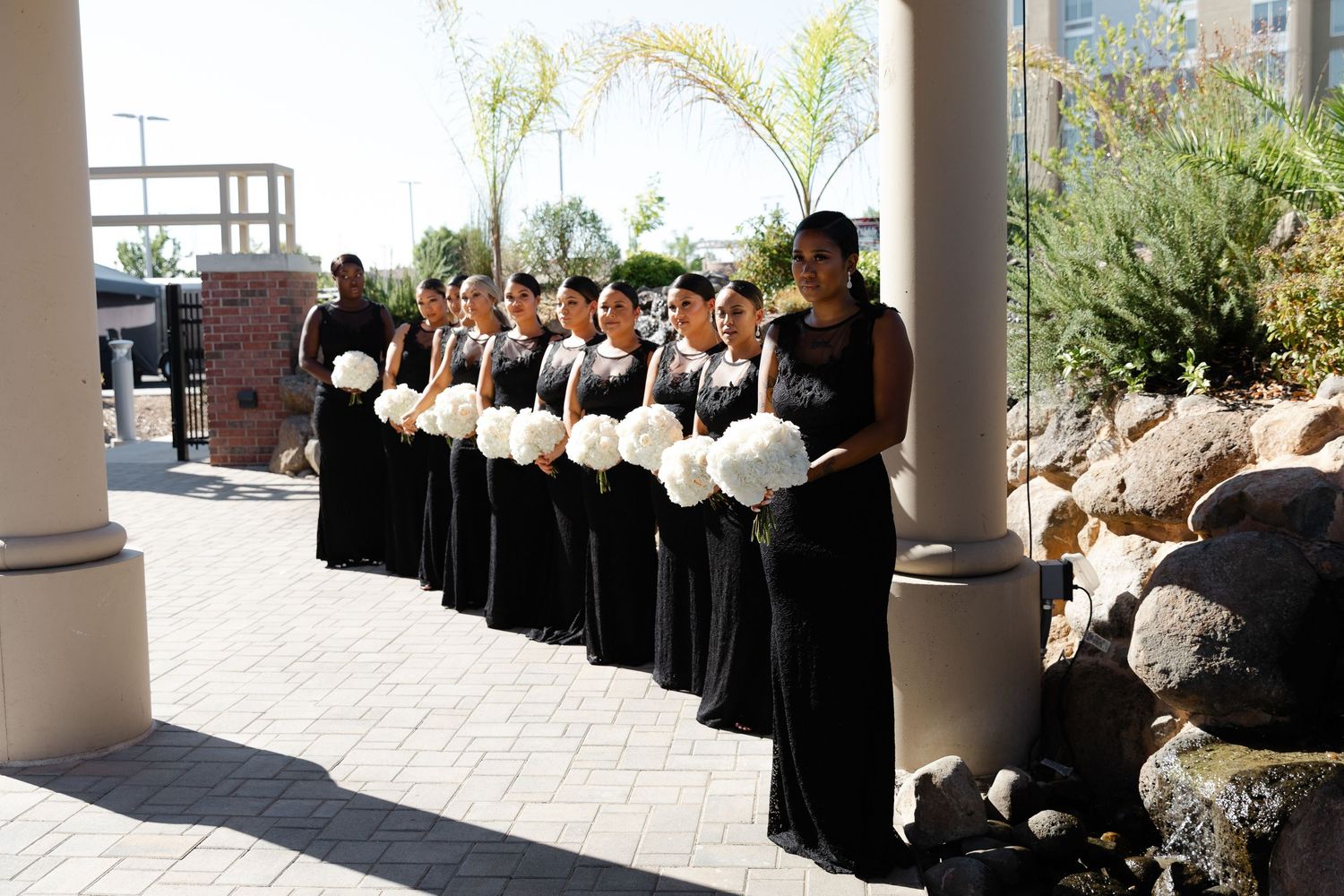 A row of people in black and white formal attire standing in a covered outdoor walkway with columns.
