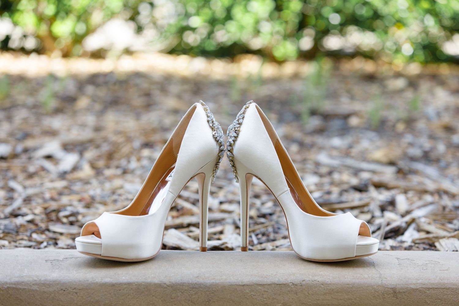 Elegant silver high heel wedding shoes with crystal embellishments photographed in an outdoor setting.