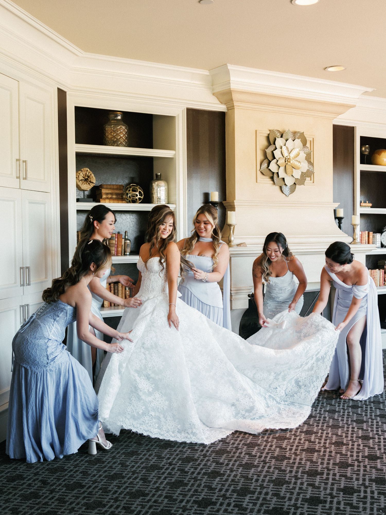 A group in light blue dresses helps arrange a white lace wedding gown in an elegant room with built-in shelving.