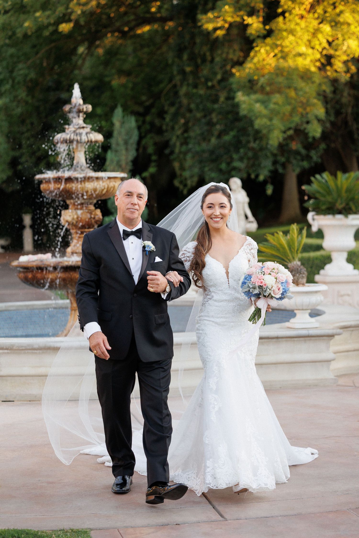 A joyful wedding couple shares a romantic moment walking together near an elegant fountain in an outdoor garden setting.
