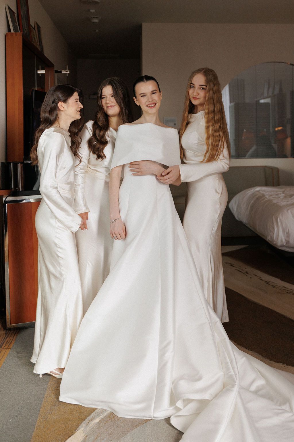 Four people in elegant white dresses stand together in a hotel room during wedding preparations.