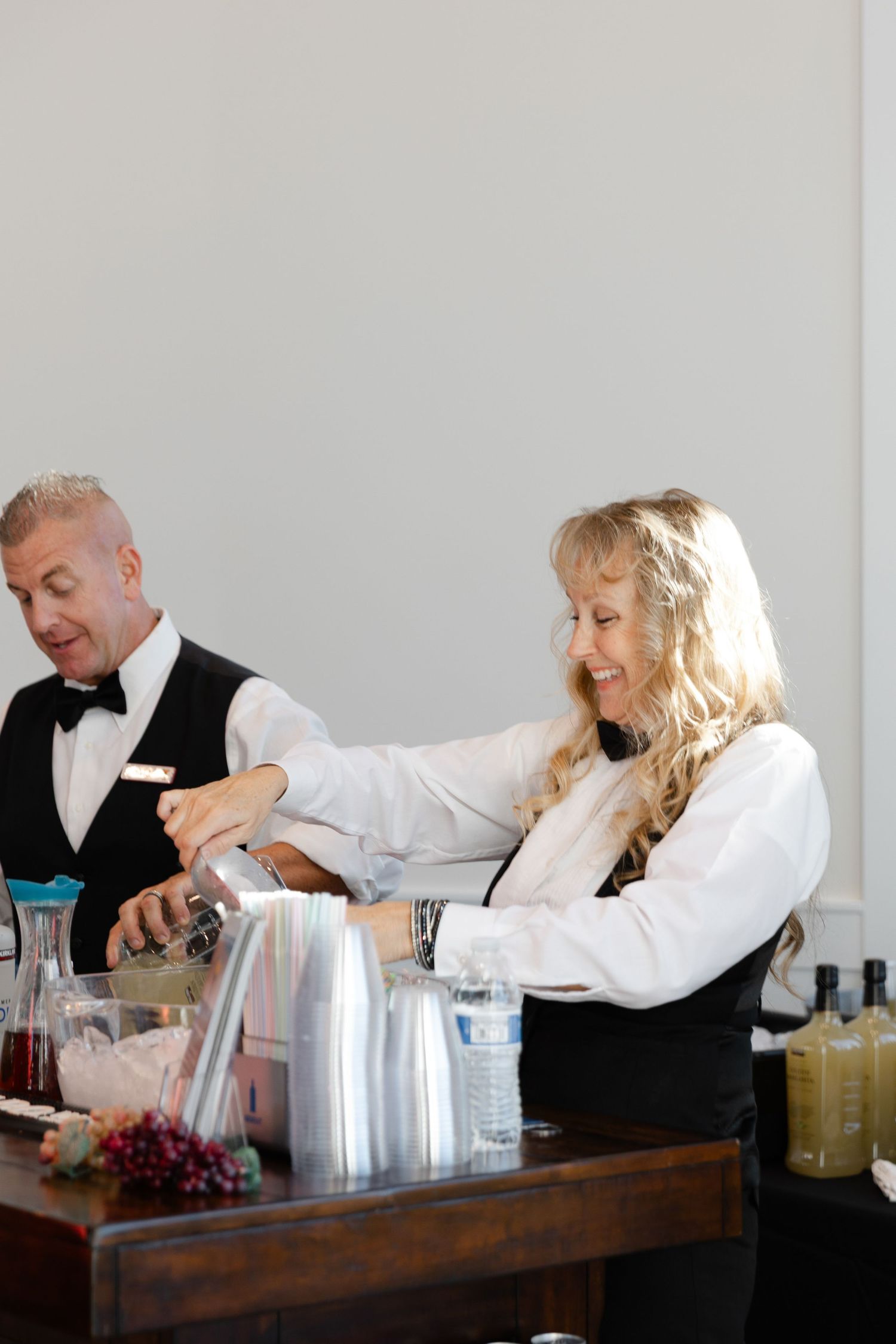 Two bartenders in white shirts and black vests mix drinks together behind a wooden bar counter.