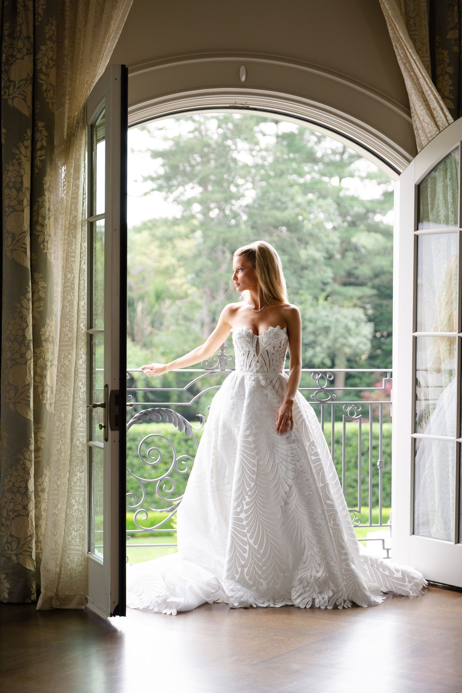 Elegant white lace wedding gown with intricate details photographed in doorway leading to balcony with ornate railings.