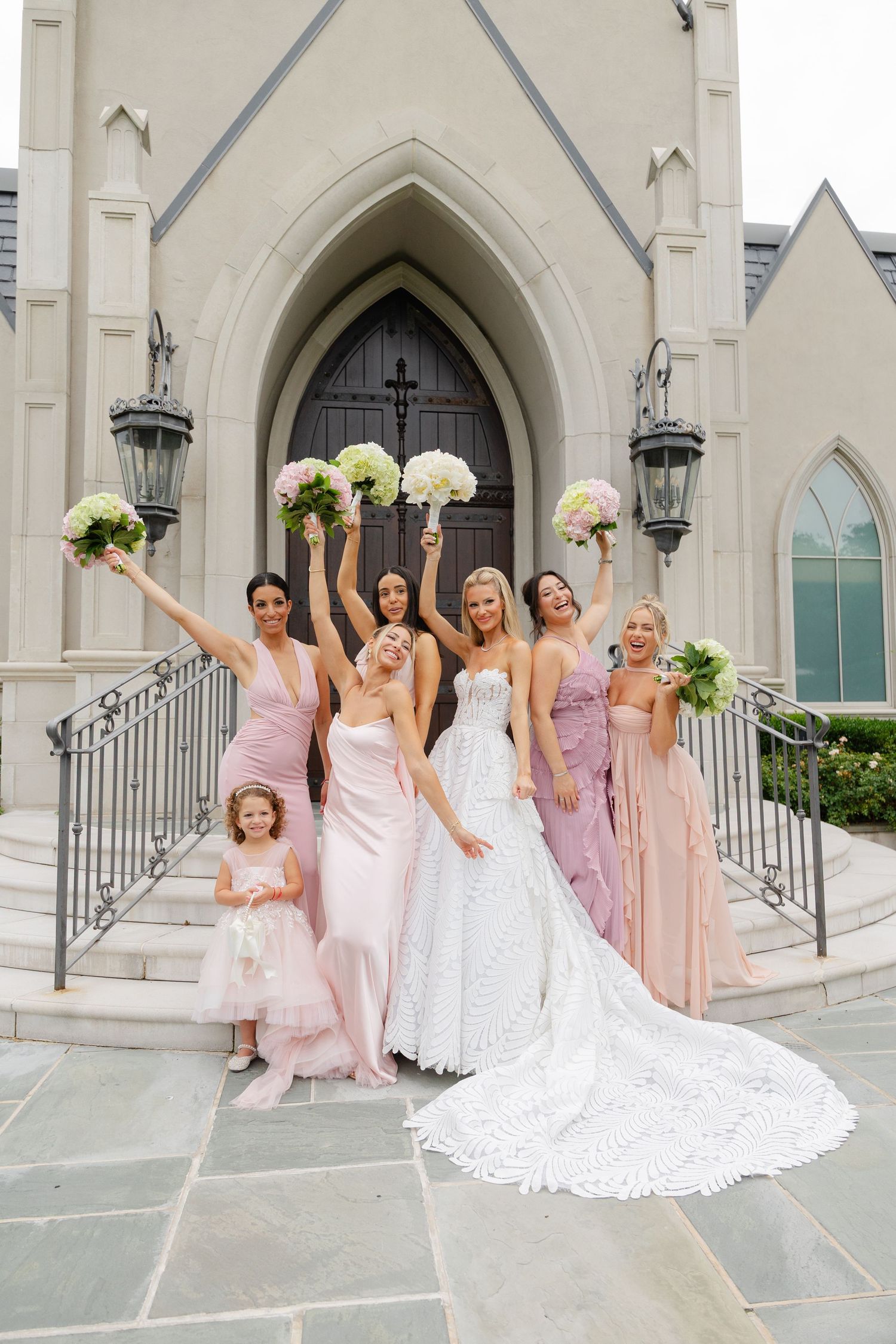 A wedding party poses on church steps with raised bouquets in front of an elegant gothic arched doorway.