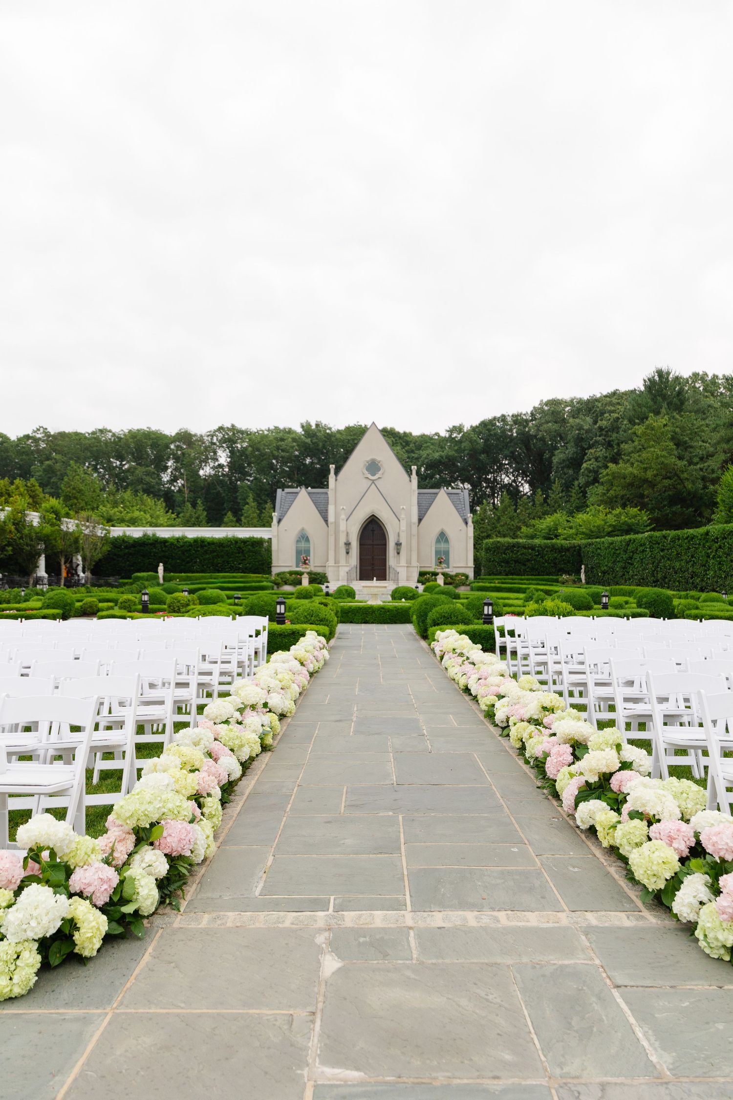 White chairs line a stone pathway leading to an elegant chapel with hydrangea floral arrangements for an outdoor wedding ceremony.