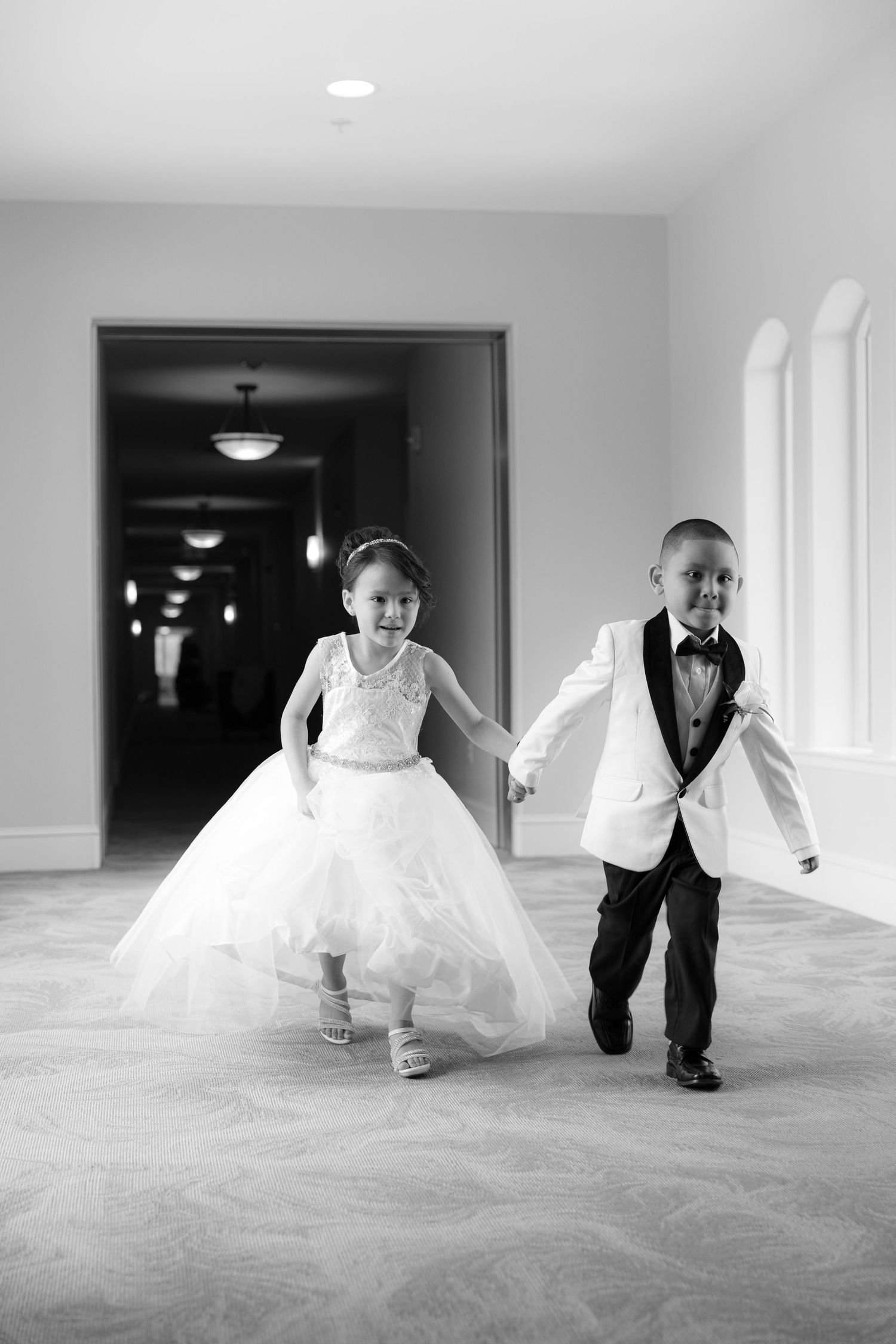 Two young children in formal wedding attire run playfully down a hallway holding hands.