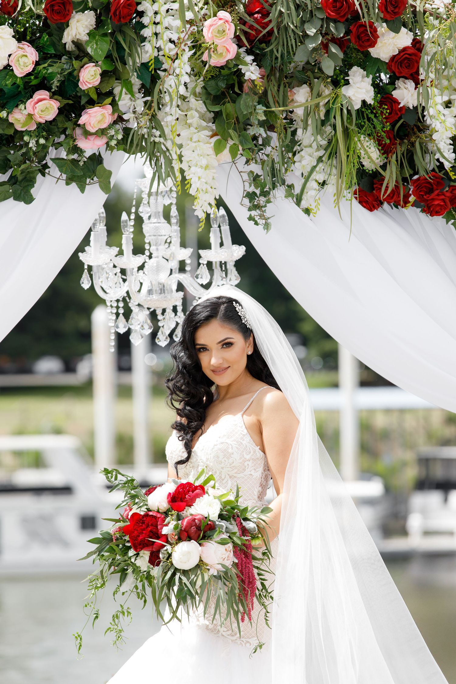 Elegant wedding arch decorated with red and blush roses, white flowers and cascading greenery with crystal chandeliers.