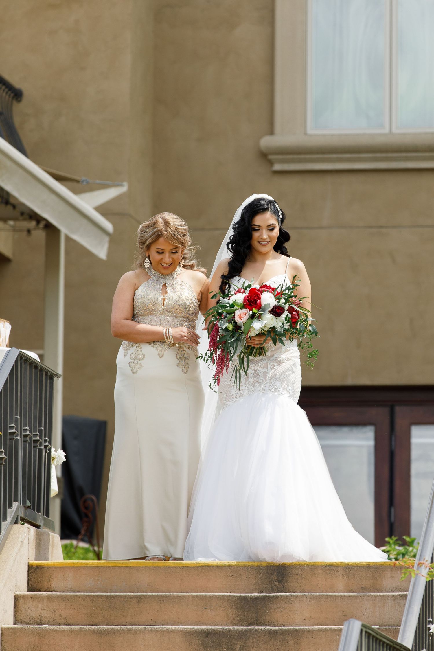 Two people in elegant white wedding gowns stand at the top of stone steps, one holding a vibrant red and white bouquet.