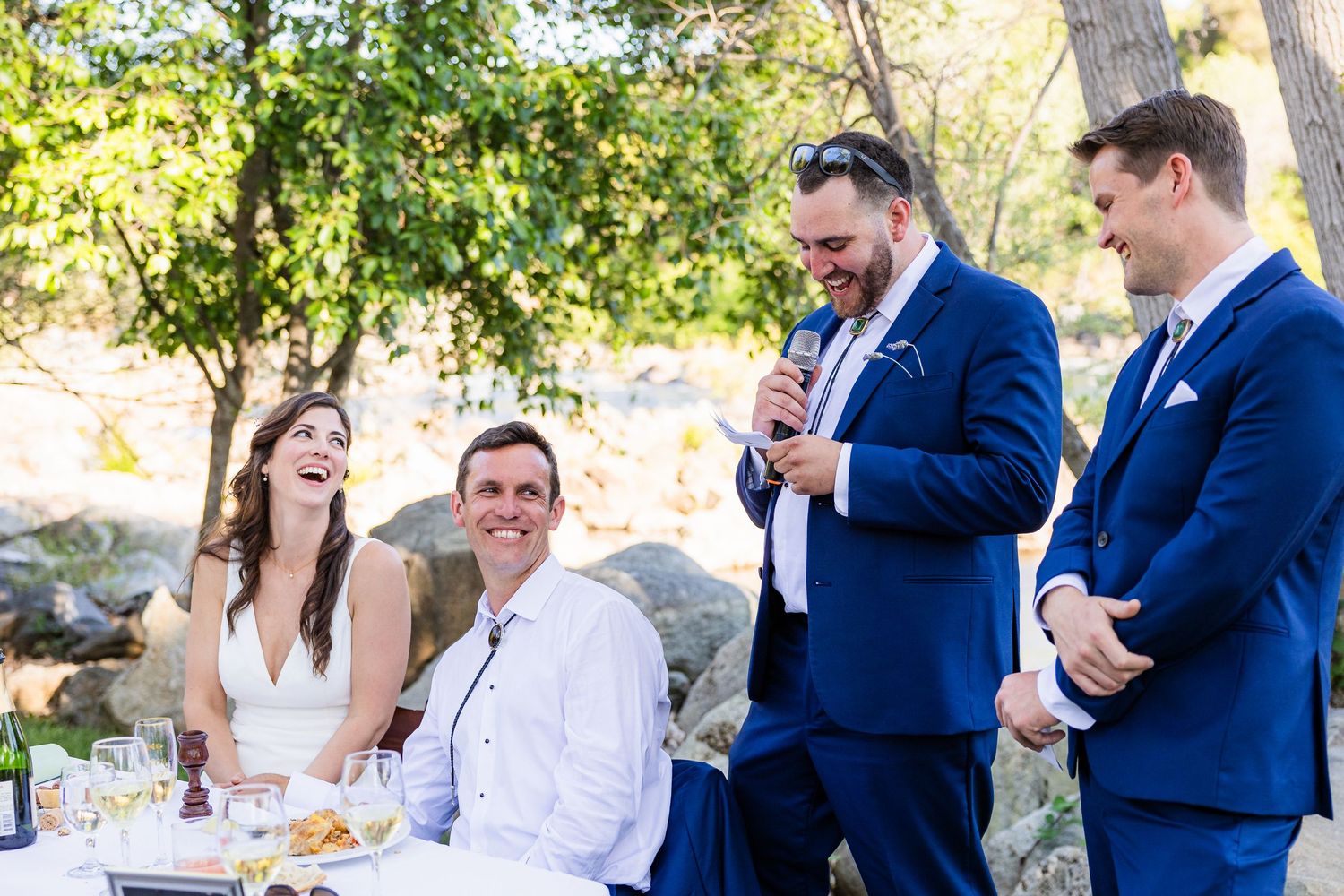 Wedding guests in navy suits and white dresses gather outdoors for a celebration under trees on a sunny day.