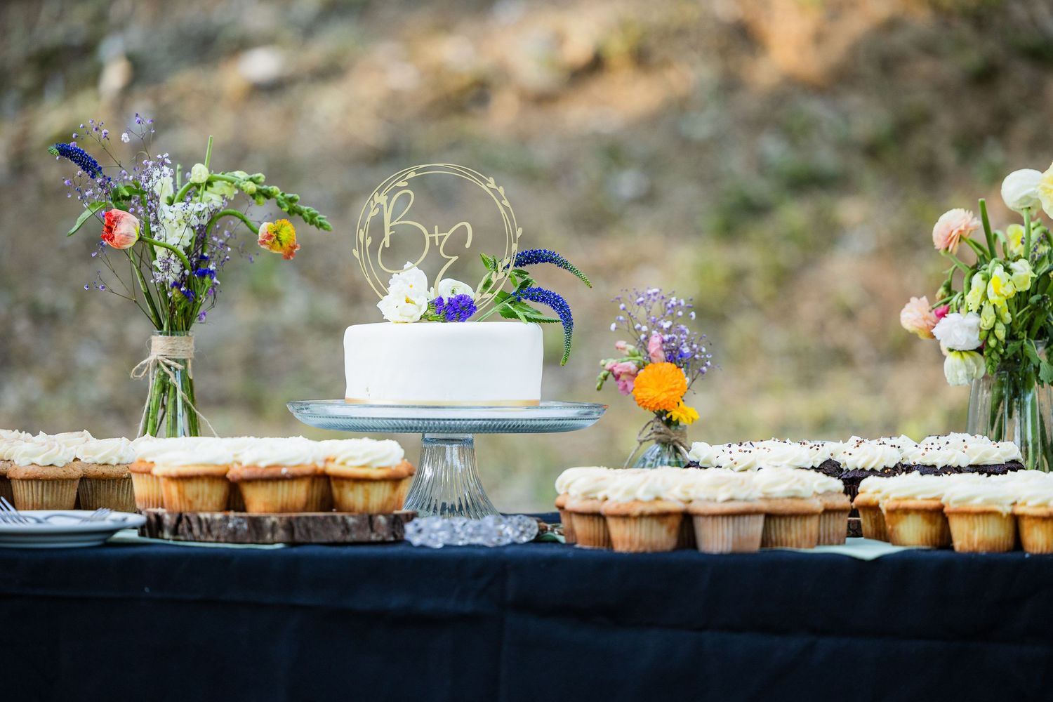 A rustic dessert table with a white wedding cake, cupcakes, and wildflower arrangements on a black tablecloth.