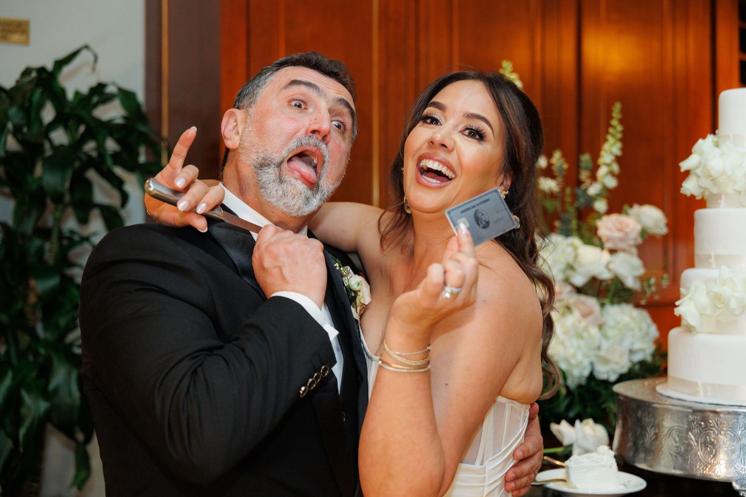 Newlywed couple shares playful moments while cutting their elegant white wedding cake decorated with flowers.