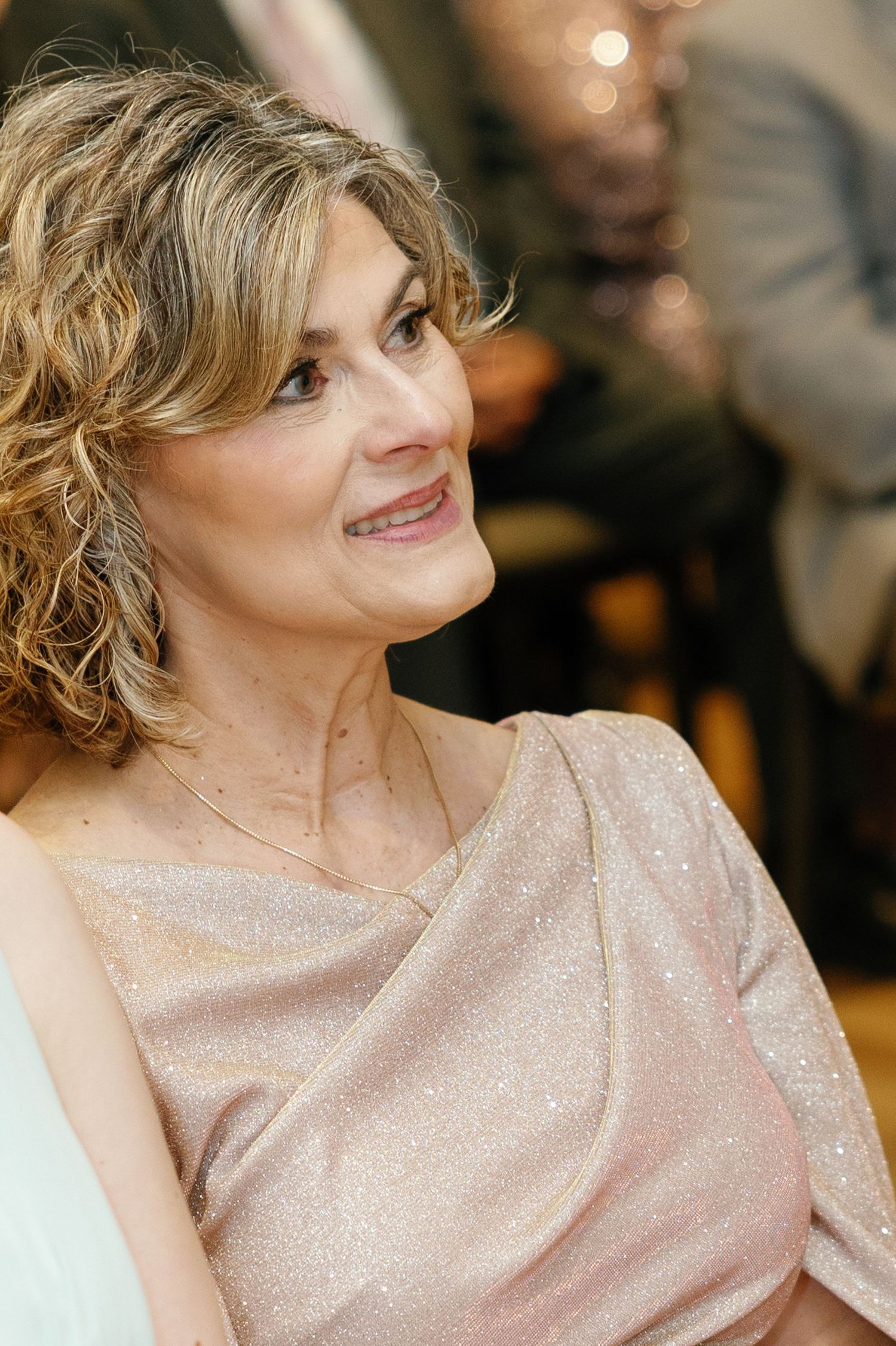 A person wearing a sparkly champagne-colored evening dress smiles warmly in an elegant indoor setting.