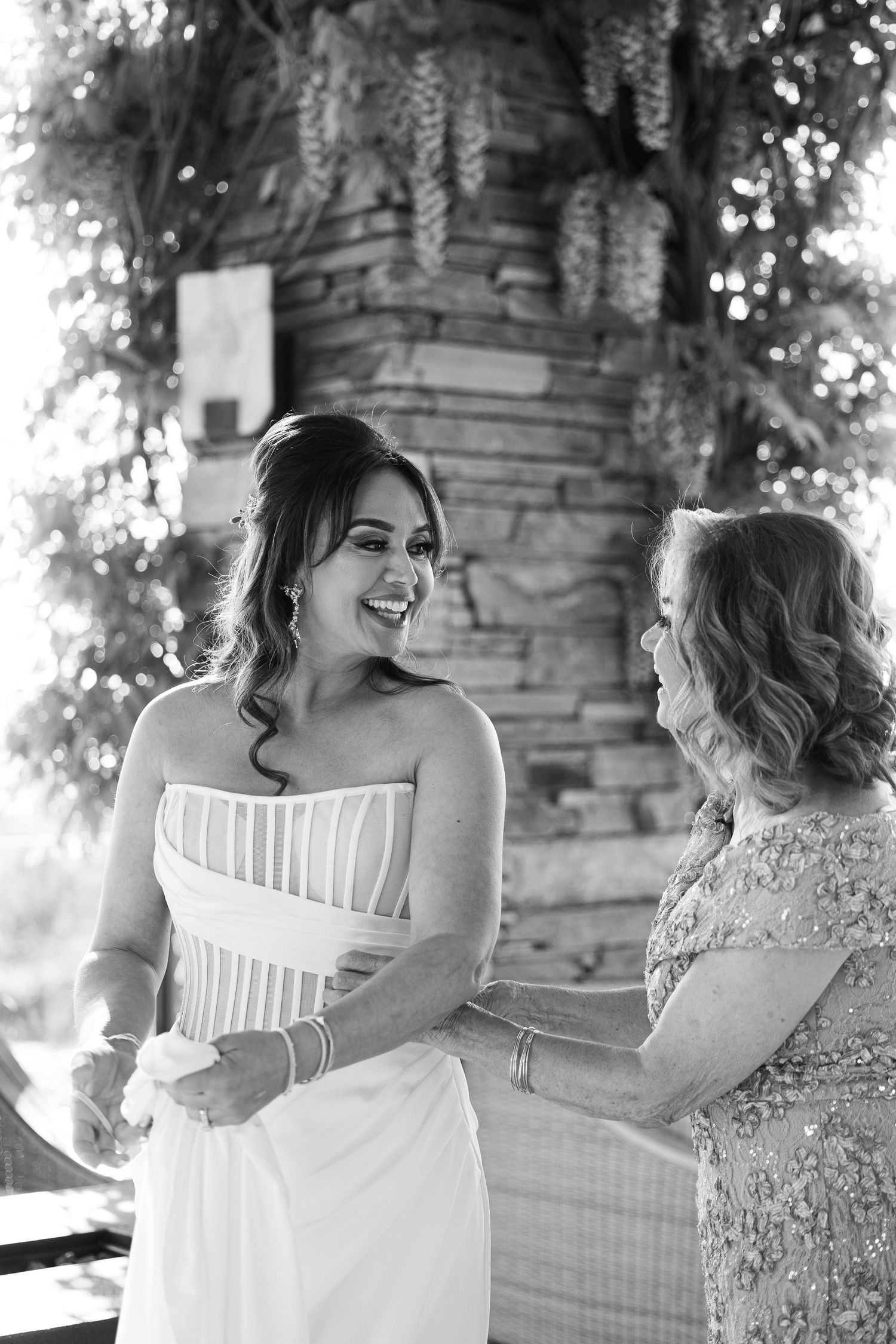 Two people in white dresses share a candid laughing moment together outdoors.