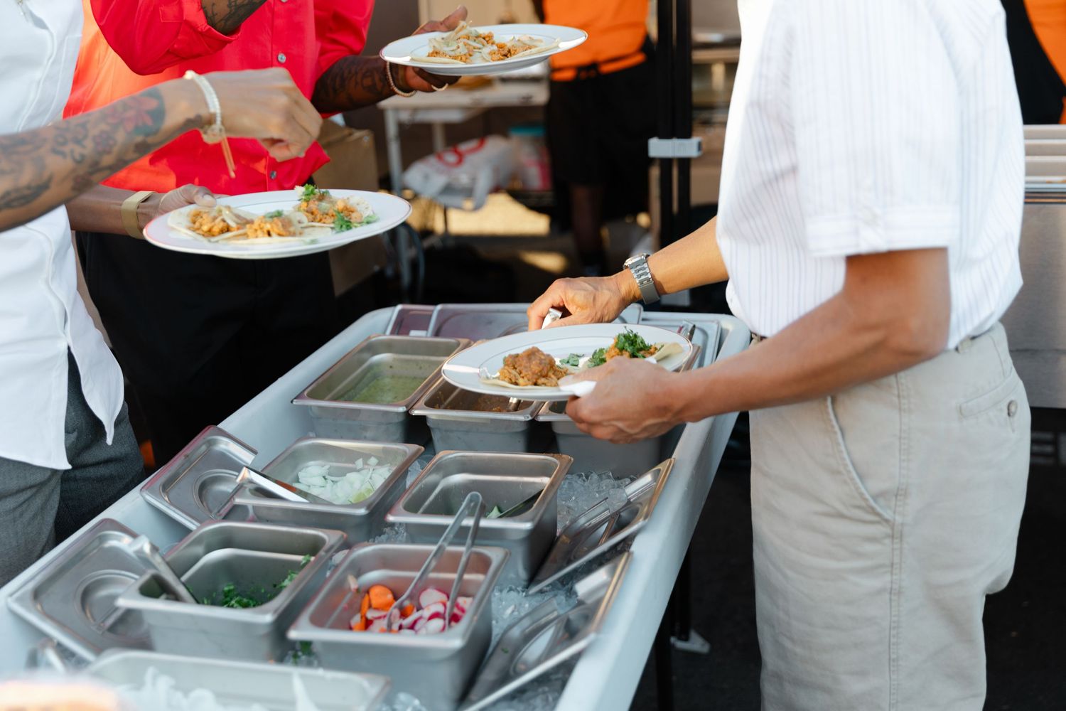 Food service workers in white uniforms serve meals from stainless steel buffet containers at a catering event.
