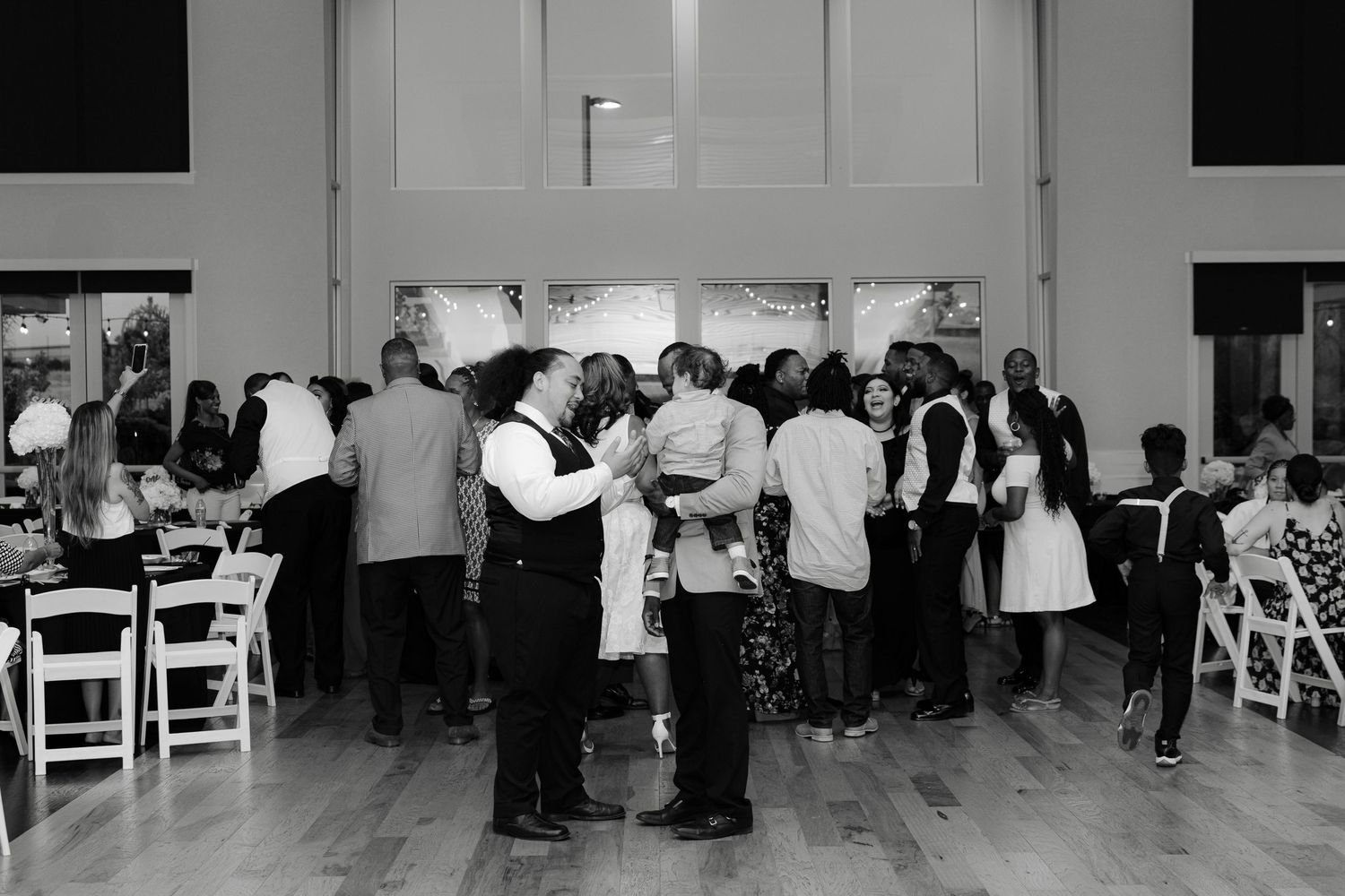 Black and white panoramic photo of wedding guests mingling in a reception hall with white chairs.