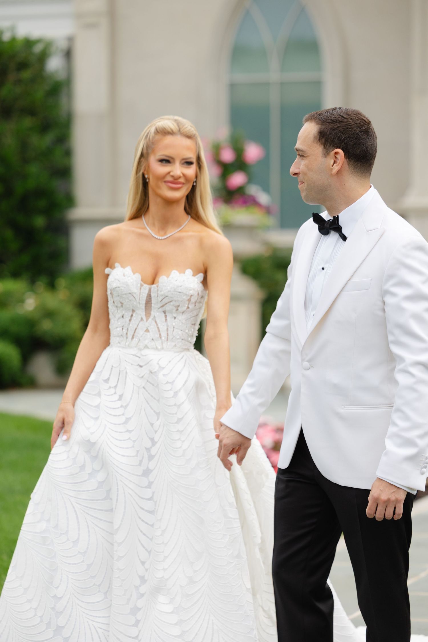 A bride in a white corset wedding dress and groom in white tuxedo jacket share intimate moments during outdoor photos.