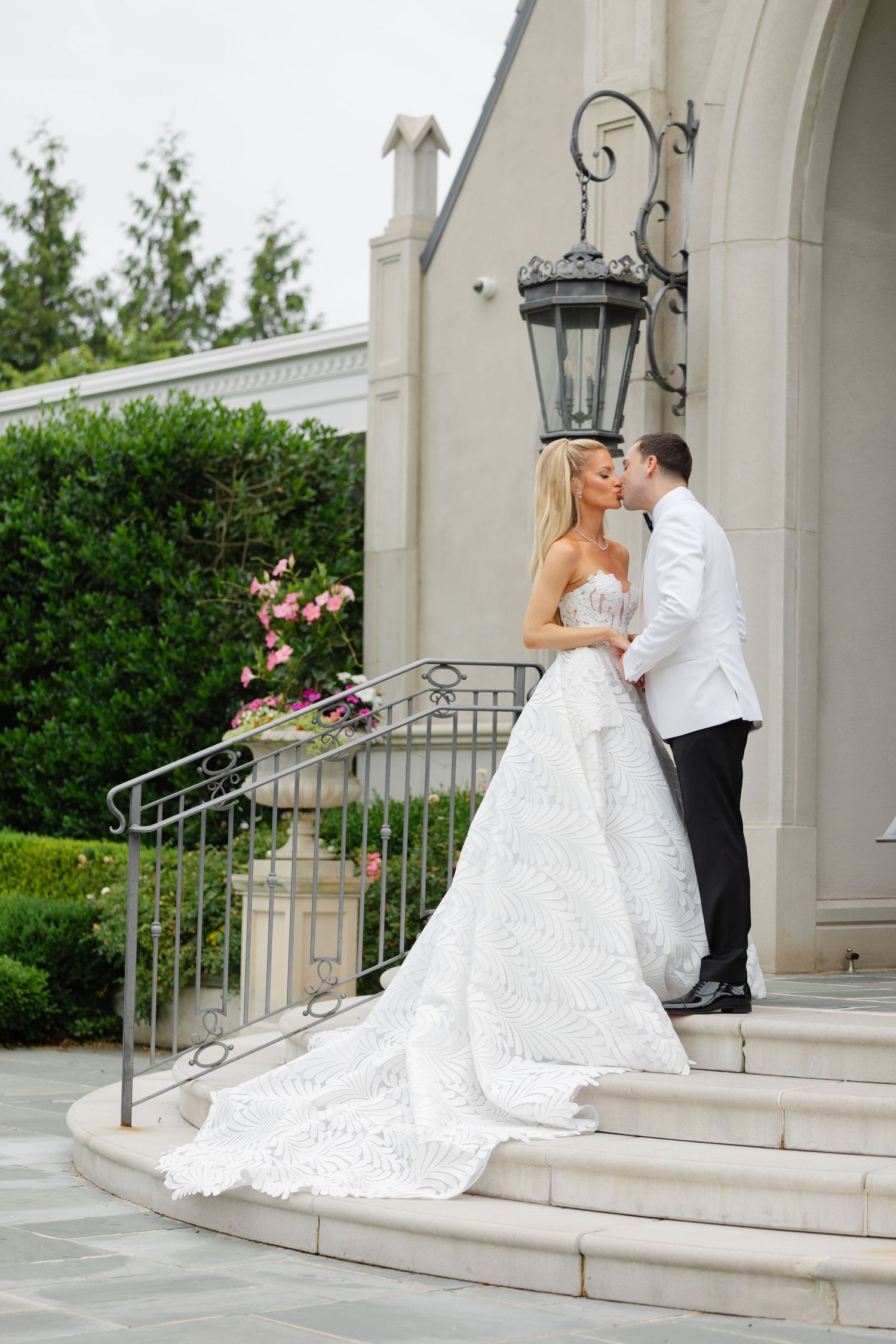 A bride in a flowing white gown and groom in a white tuxedo share a romantic kiss on elegant stone steps.