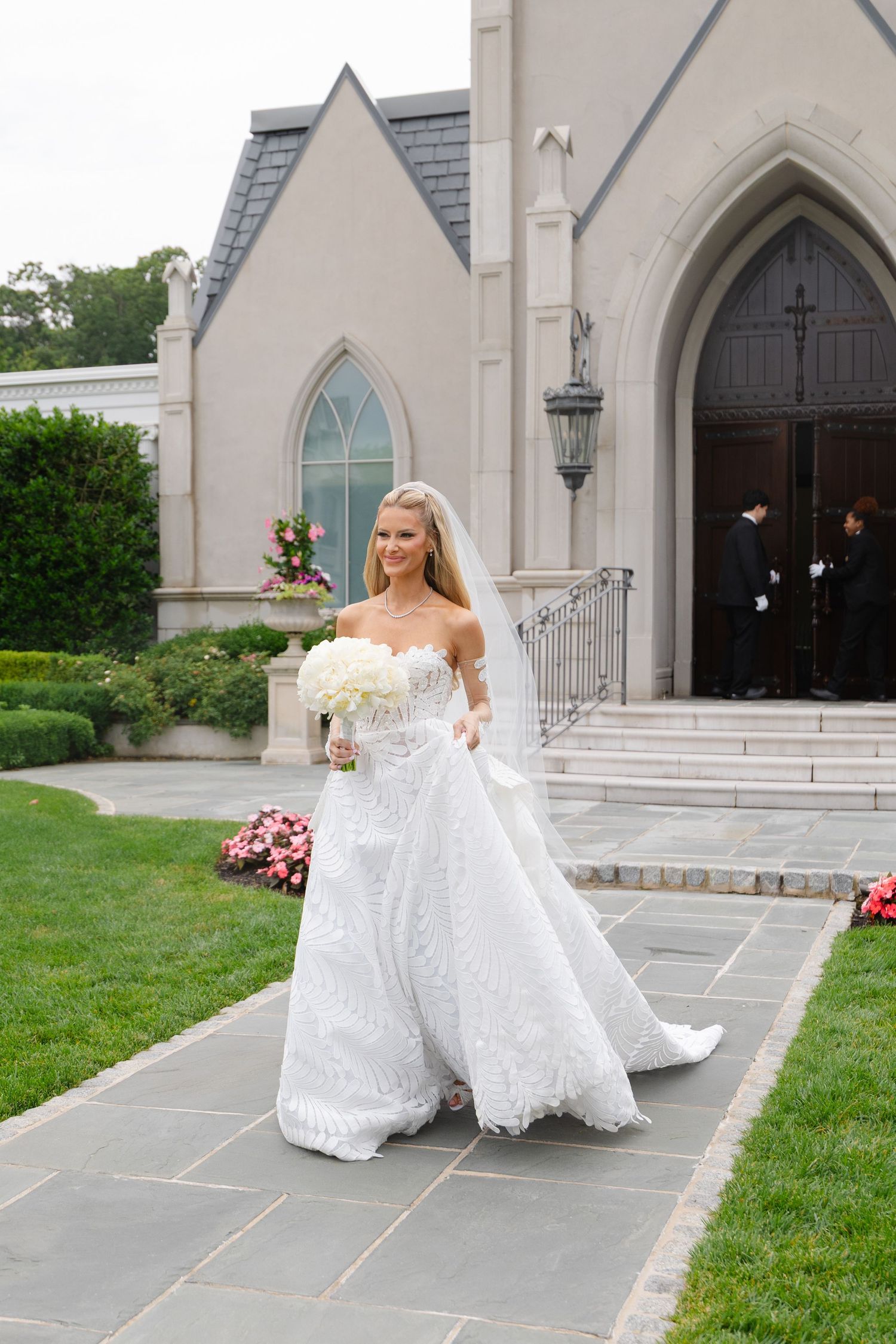 Smiling bride in strapless lace wedding gown with veil holding white bouquet walking outside a stone chapel.