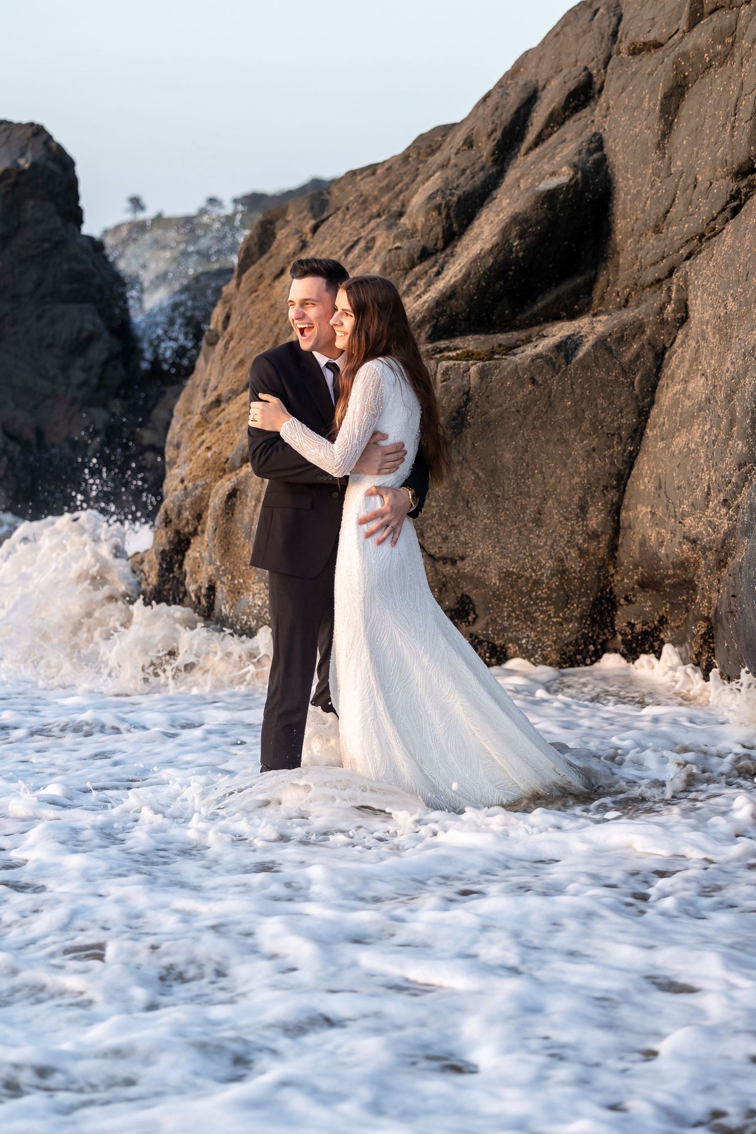 Couple in wedding attire embraces near rocky coastline with crashing waves.