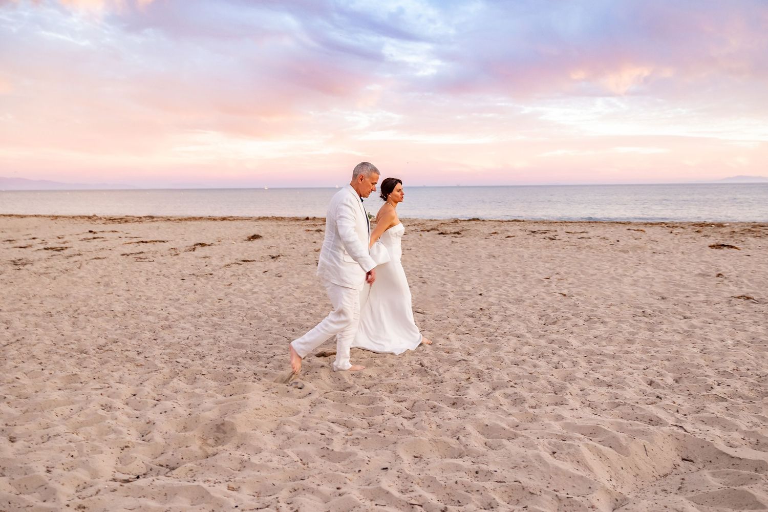 Couple walks along sandy beach at sunset in wedding attire.