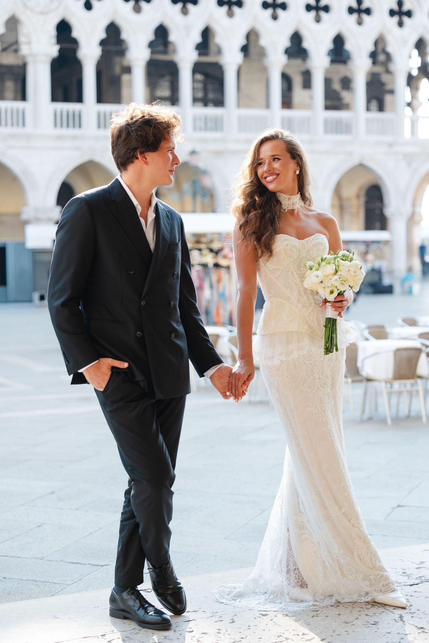 A couple in wedding attire shares a romantic moment in a beautiful outdoor courtyard with white stone architecture.