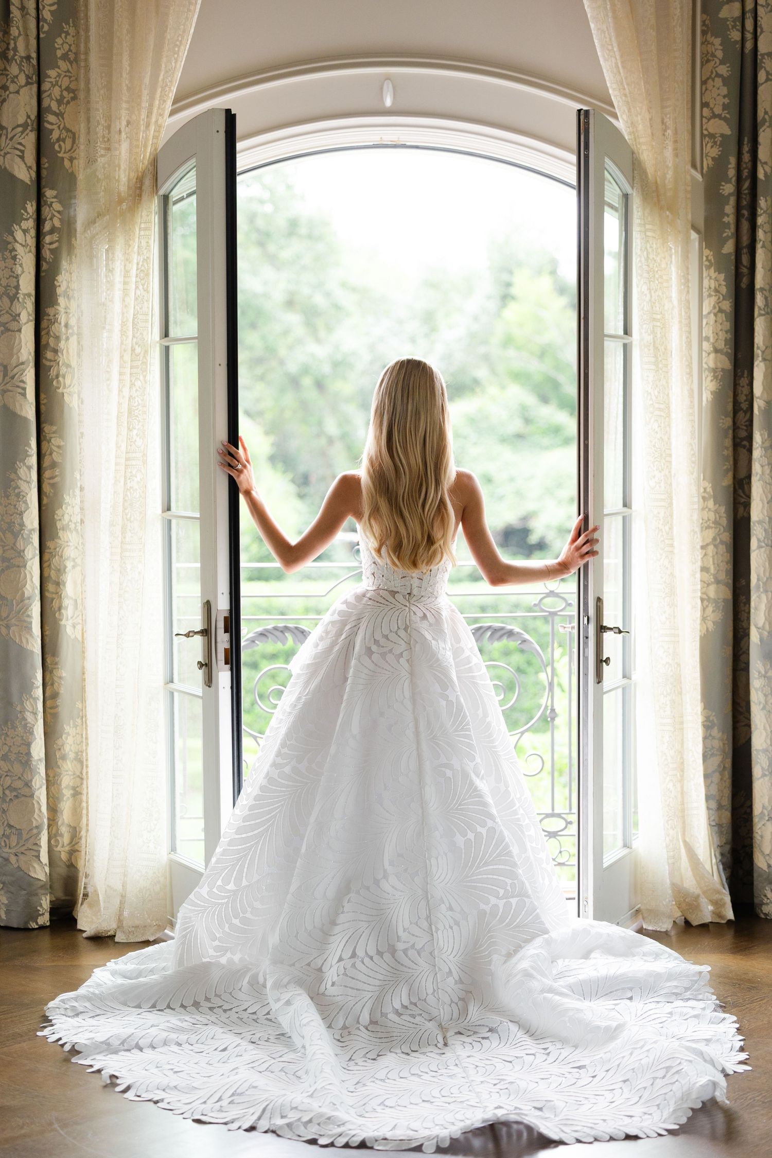 Bride in elegant white ballgown with lace train stands at open French doors overlooking lush garden.