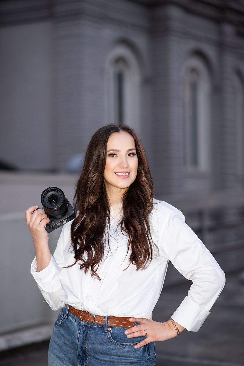 Young woman photographer smiling in white blouse and jeans, holding a DSLR camera outdoors near a gray stone building.