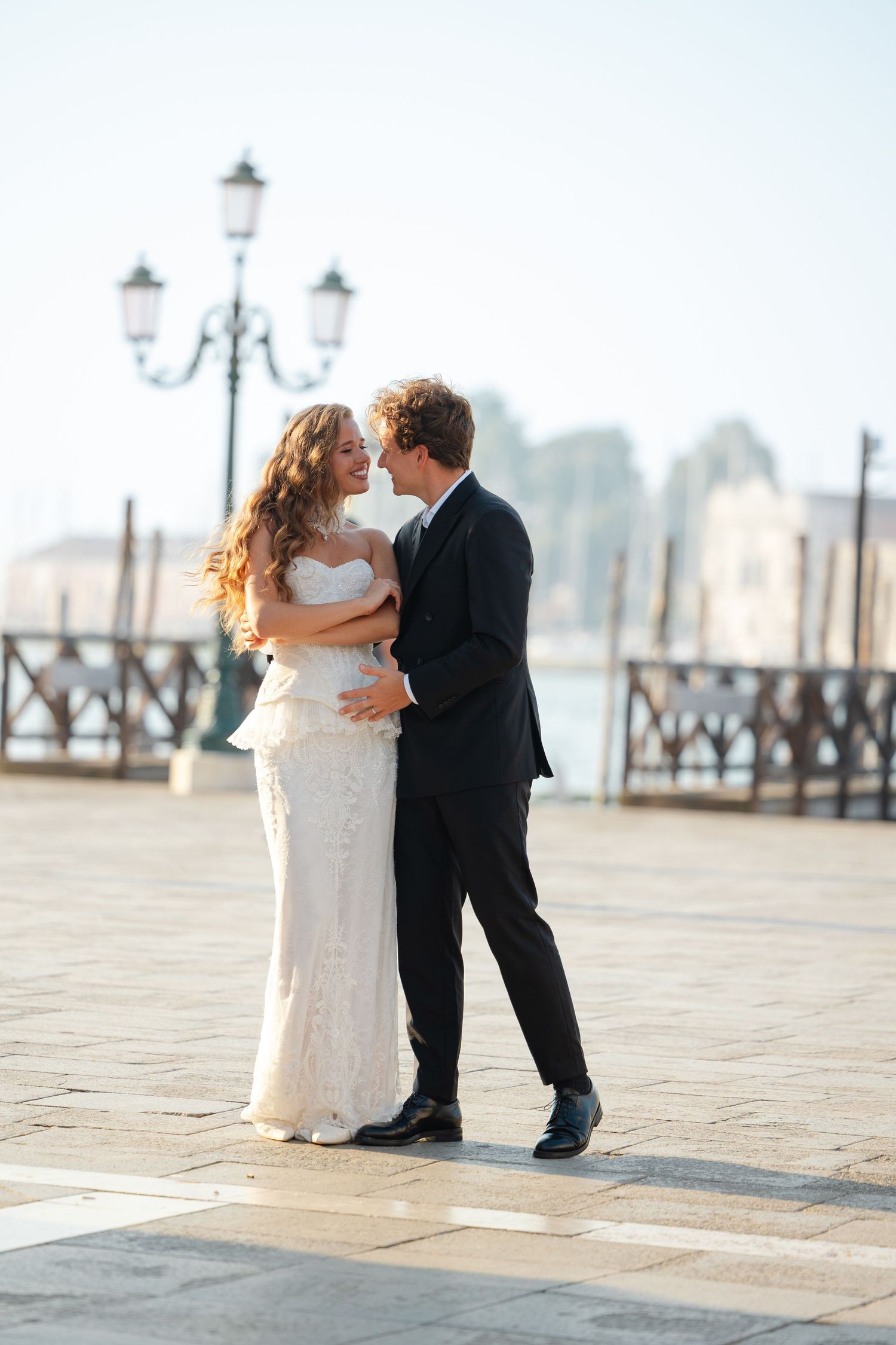 A romantic wedding couple embraces on a Venice boardwalk with ornate street lamps and architecture in the background.
