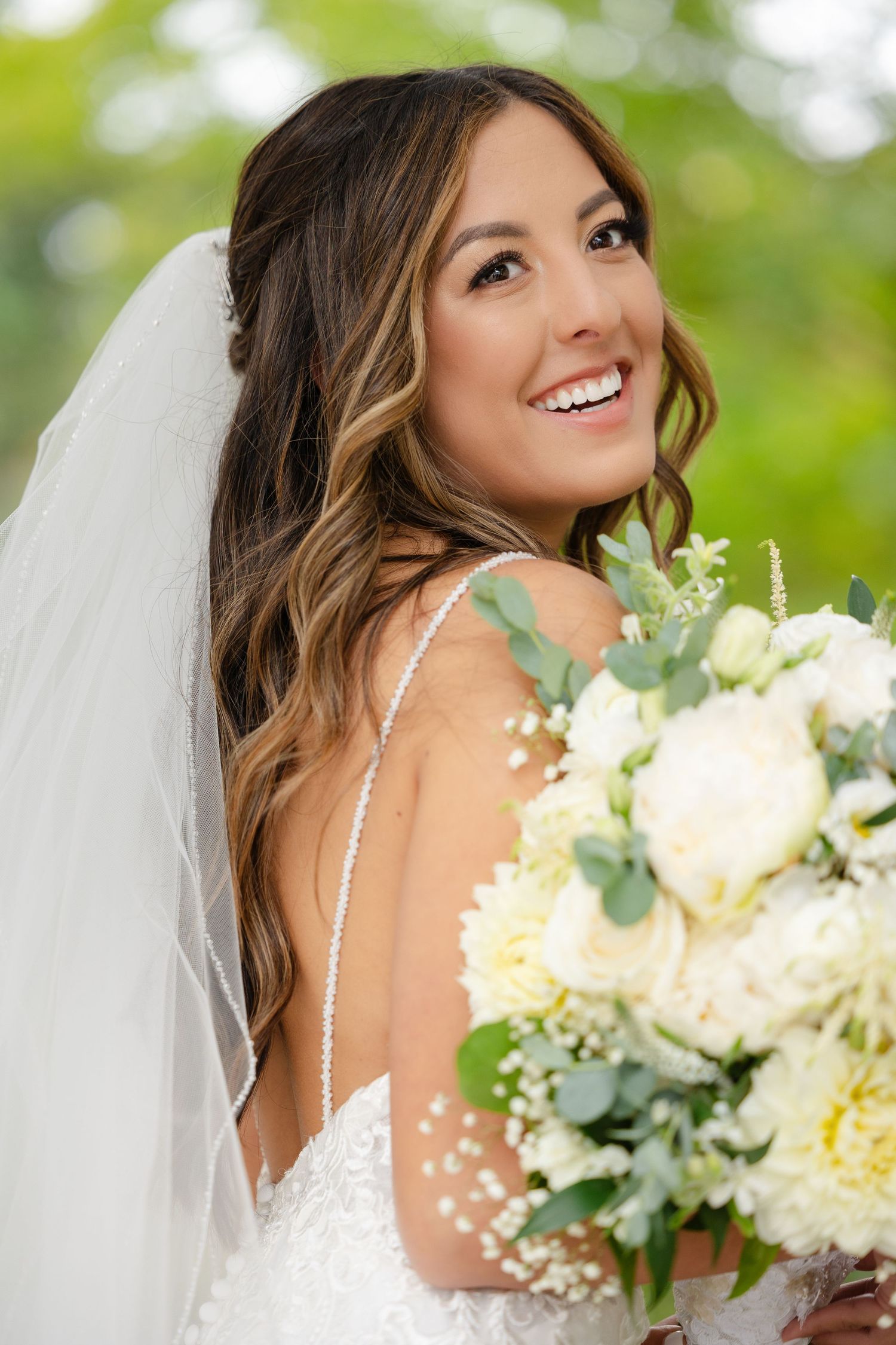 A bride in a white dress smiles while holding a beautiful bouquet of white flowers and eucalyptus against a blurred green background.