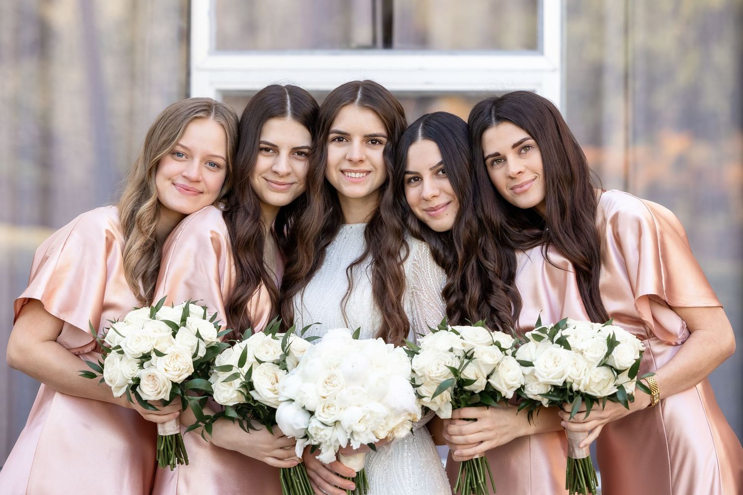 Bridesmaids in blush pink dresses hold white floral bouquets while smiling.