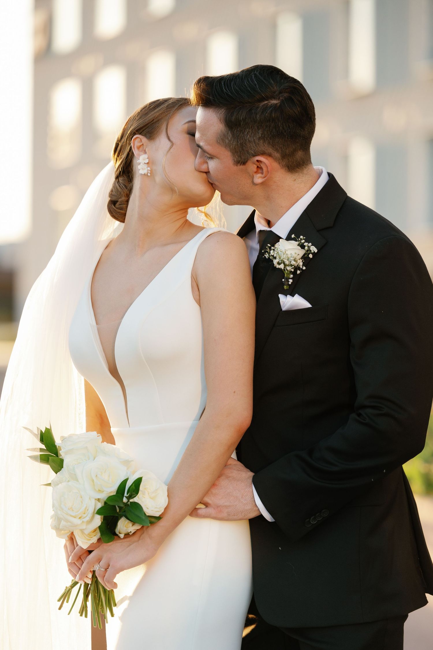 Romantic wedding couple sharing a tender kiss in golden sunlight, bride in elegant white gown holding white bouquet.