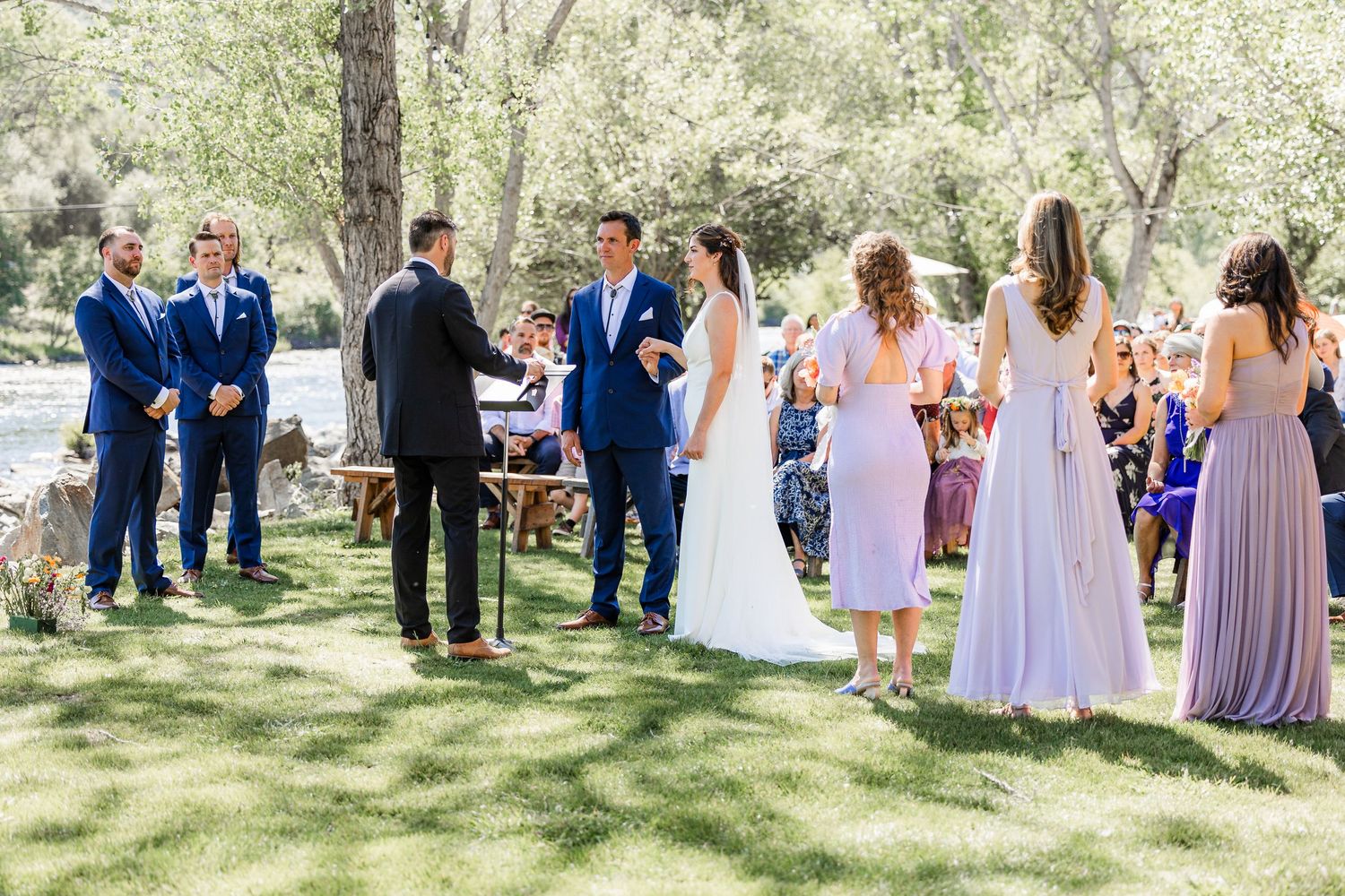 Wedding party in blue suits and lavender dresses stands on grass during an outdoor ceremony.