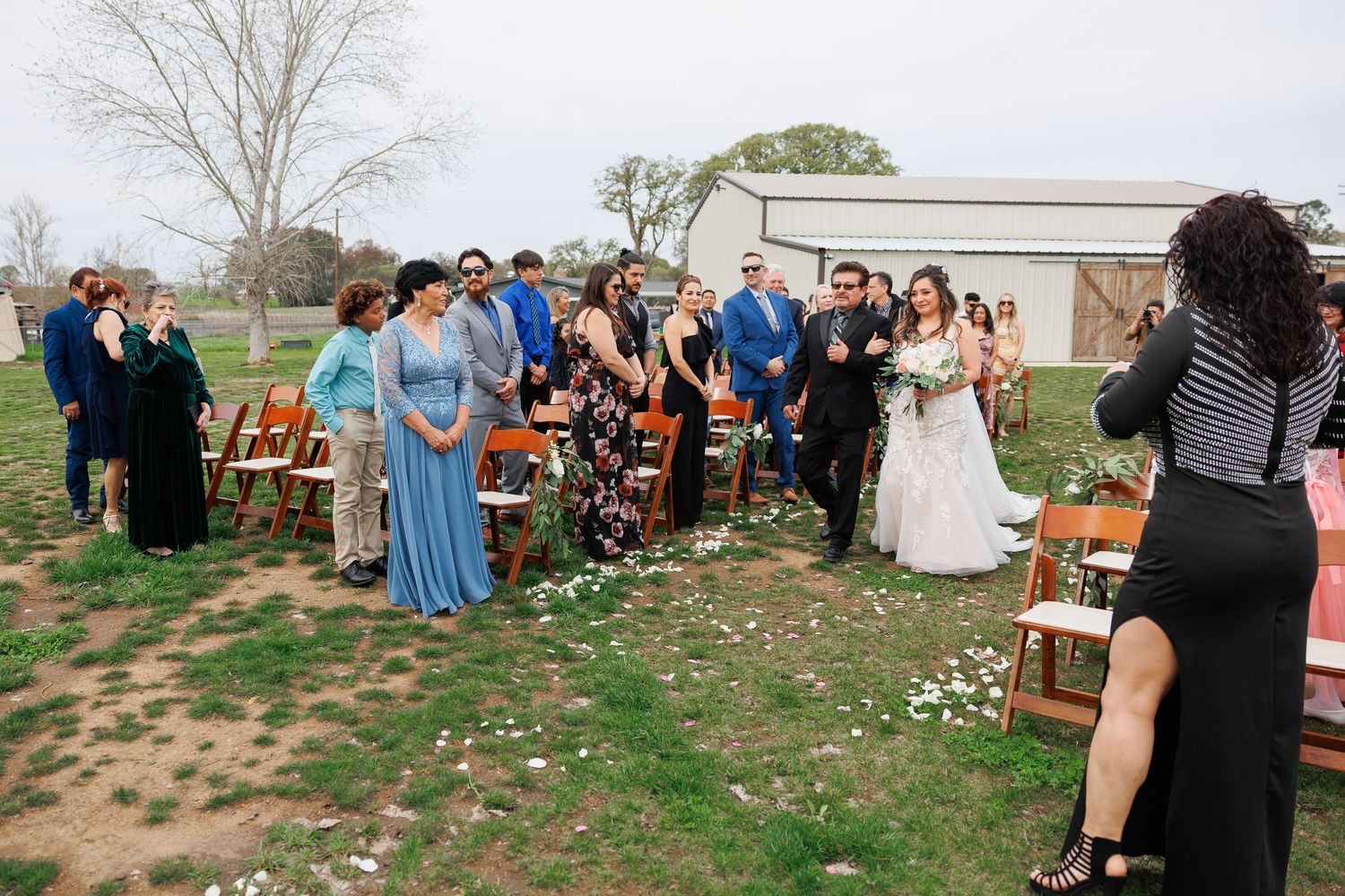 A group of wedding guests gather outdoors for a casual ceremony with wooden chairs and natural landscaping.