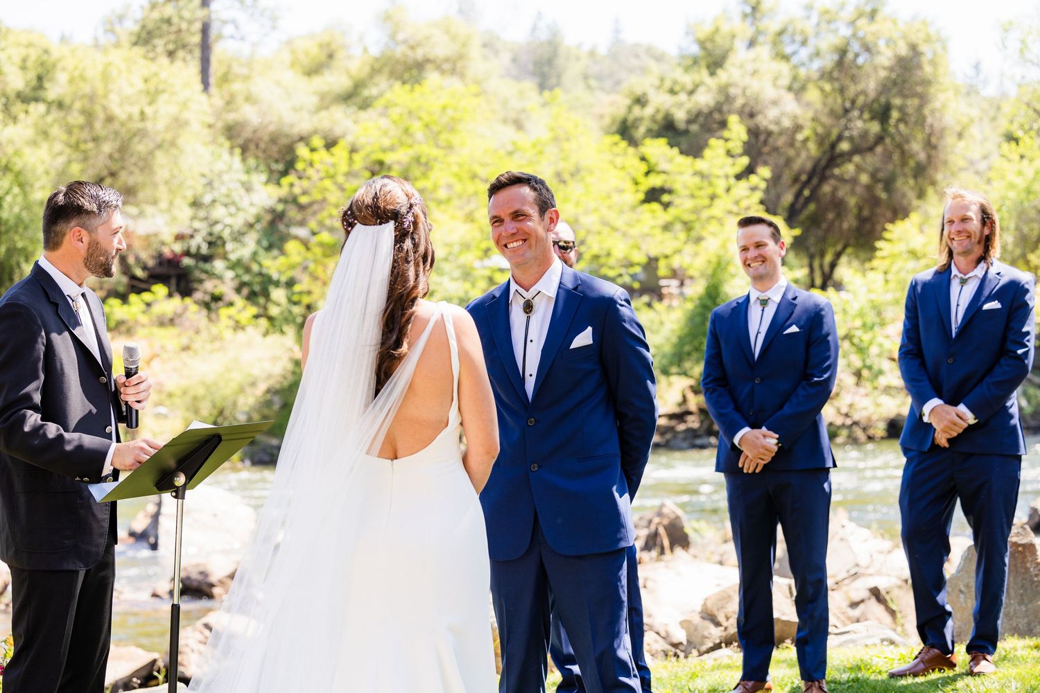 Wedding party in navy blue suits and formal attire poses outdoors in a sunny garden setting.