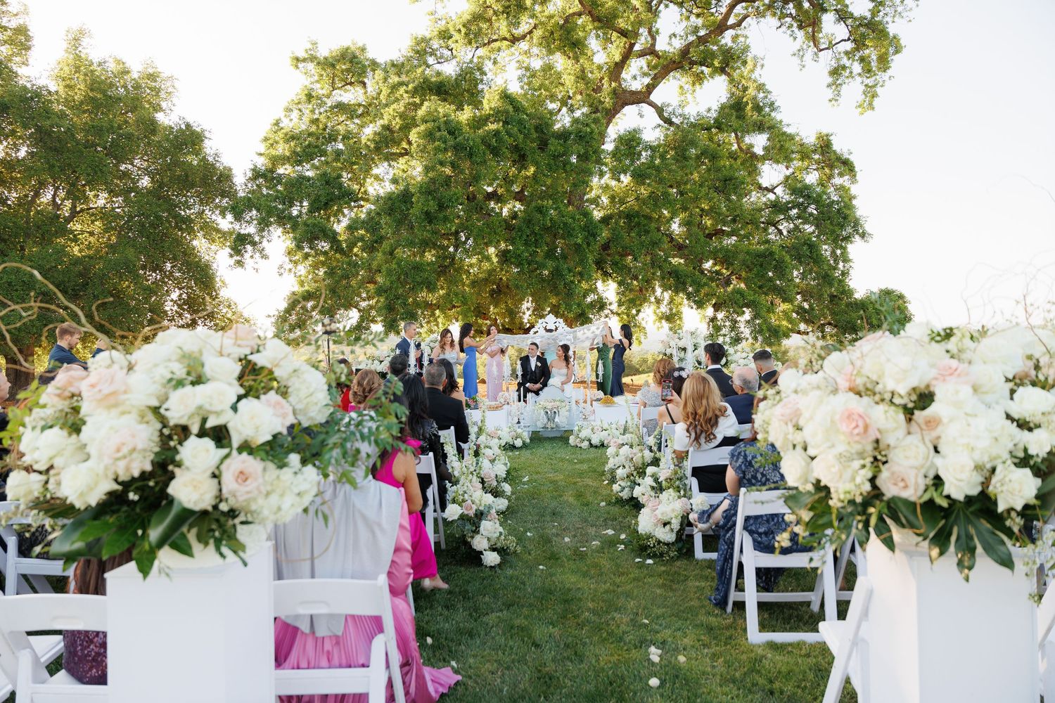 White floral wedding arch arrangements decorate an outdoor ceremony space with trees in the background.