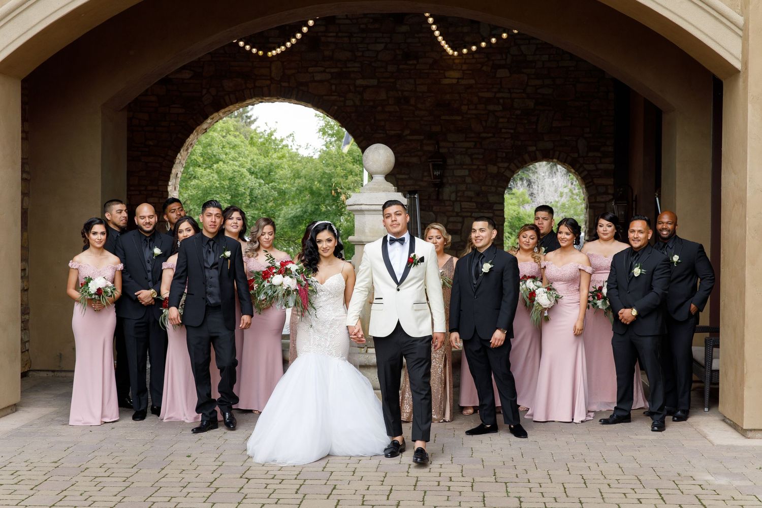 Wedding party in formal attire poses under stone archways during outdoor ceremony.