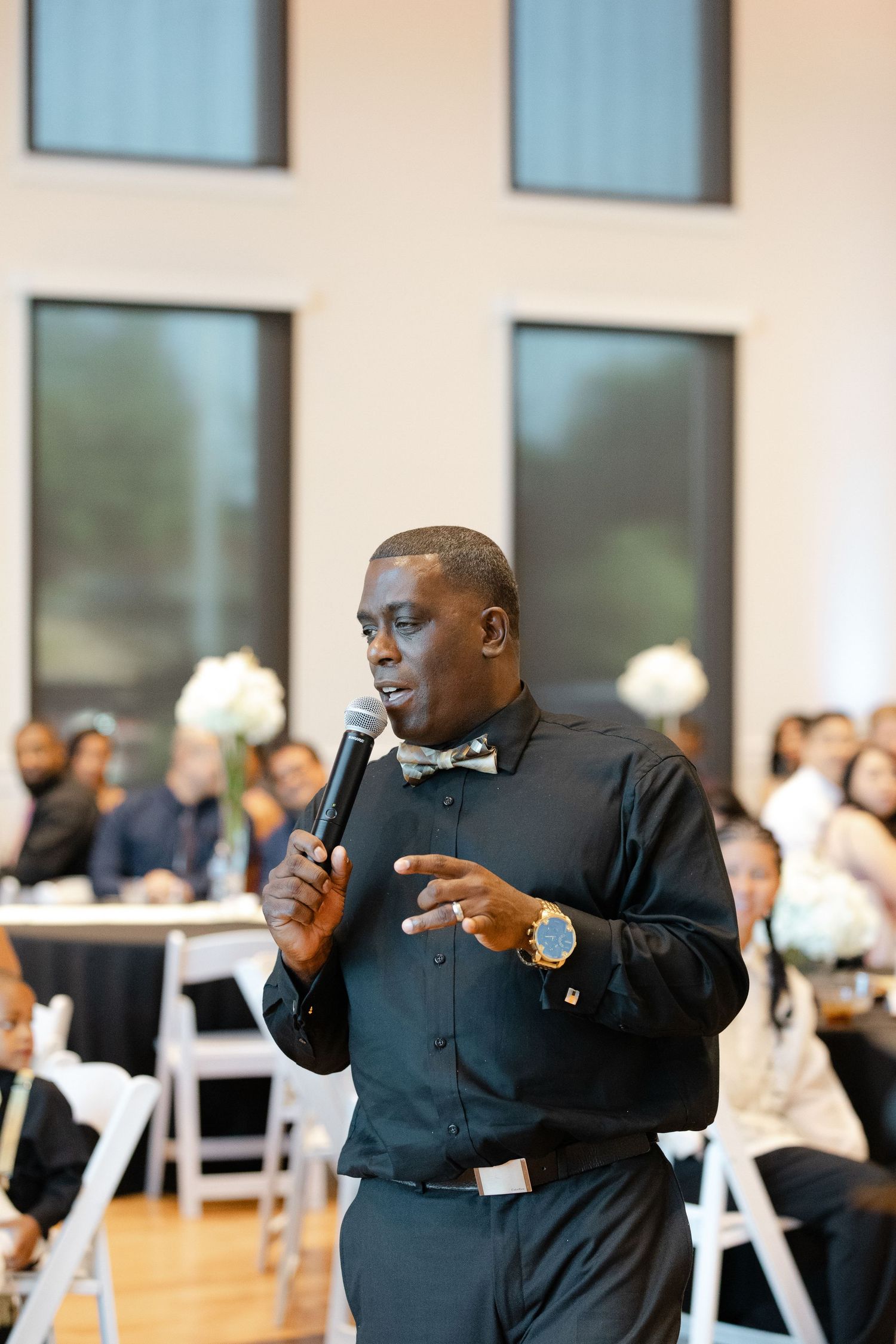 A speaker in a black suit gestures energetically while giving a presentation in front of a bright windowed room.