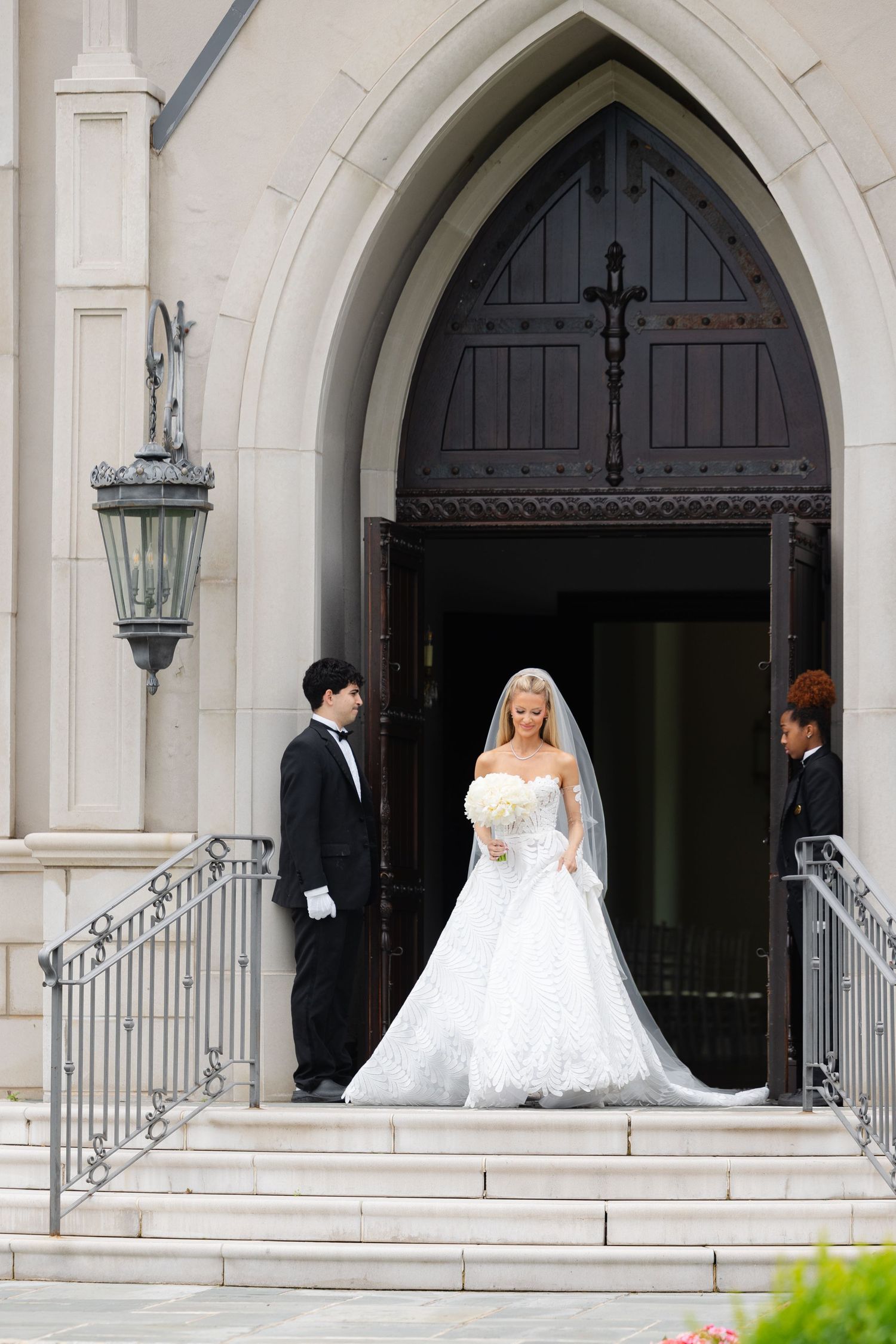 A wedding party stands in the arched doorway of a classic church with ornate architecture and iron railings.