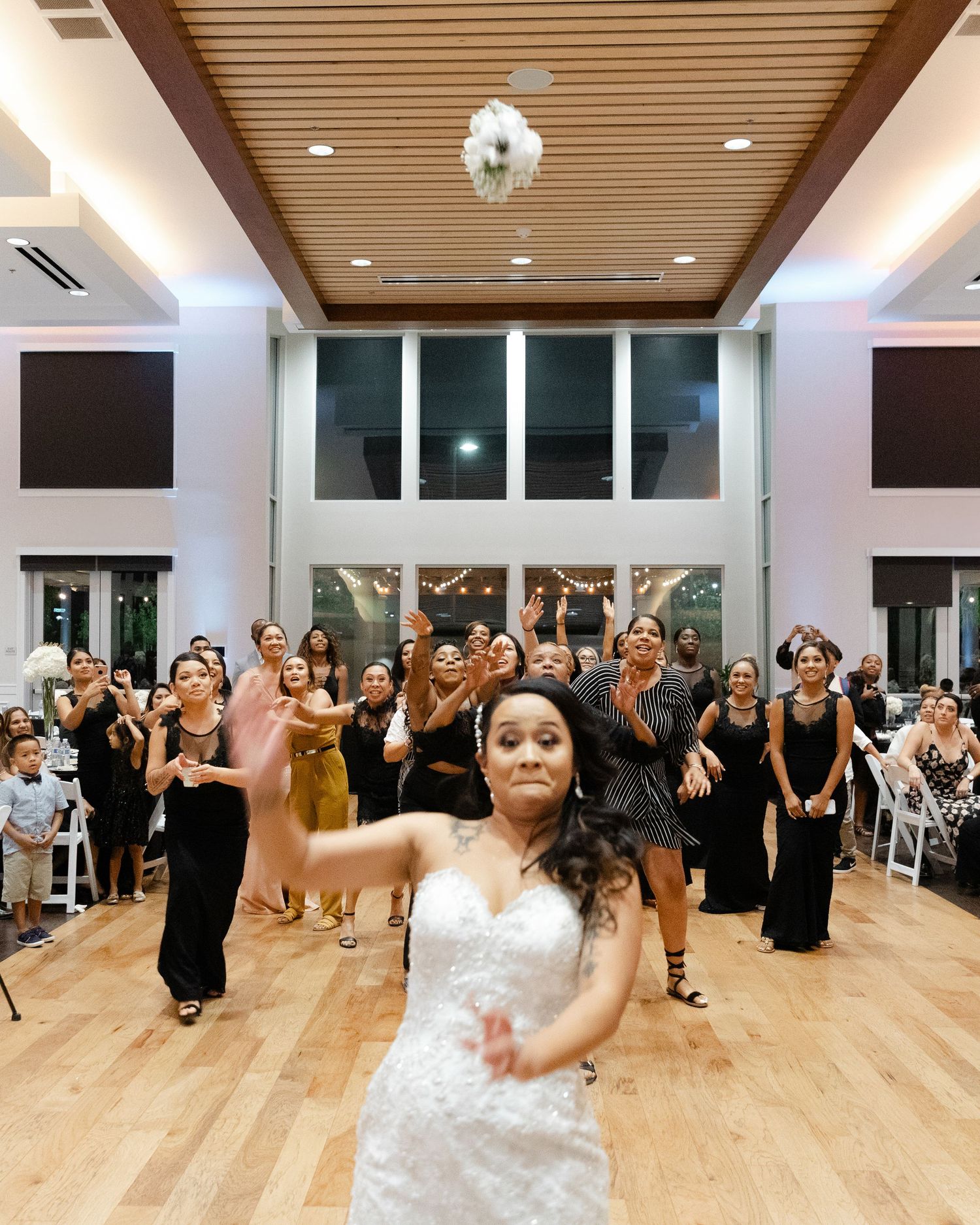 A bride tosses a bouquet during a wedding reception as guests in black dresses watch from behind on a wooden dance floor.