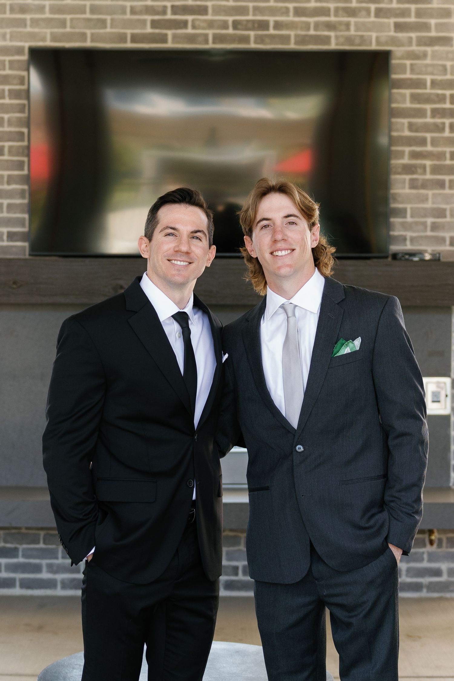 Two men in black suits and ties smile and pose together against a brick wall at a formal event.