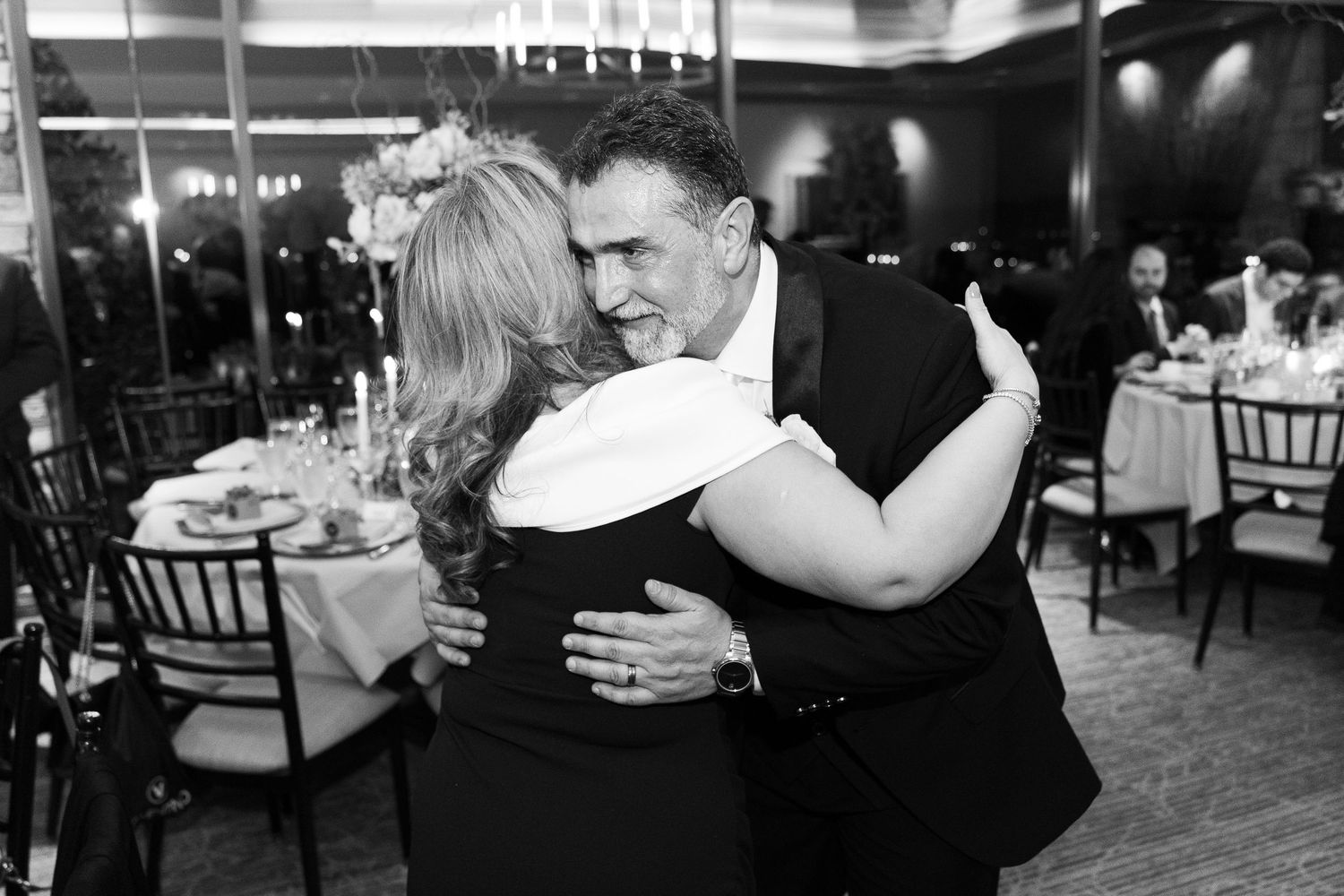 A black and white photo of a couple dancing together at an elegant reception venue with tables and chairs visible.