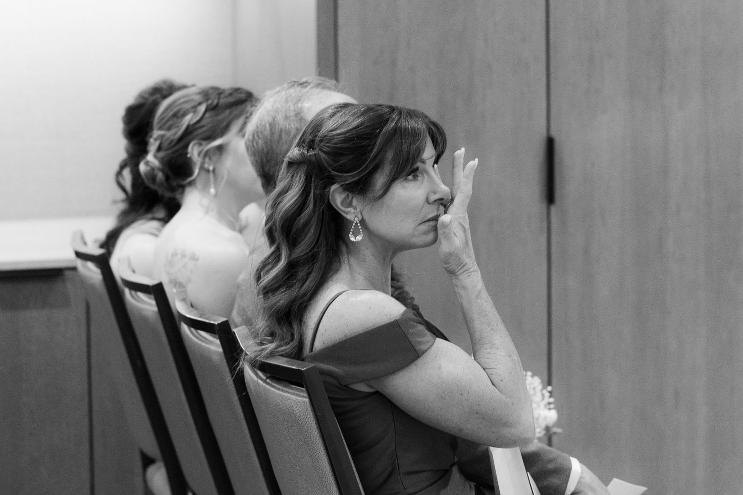A black and white photo shows people seated in wooden chairs while watching something off-screen.