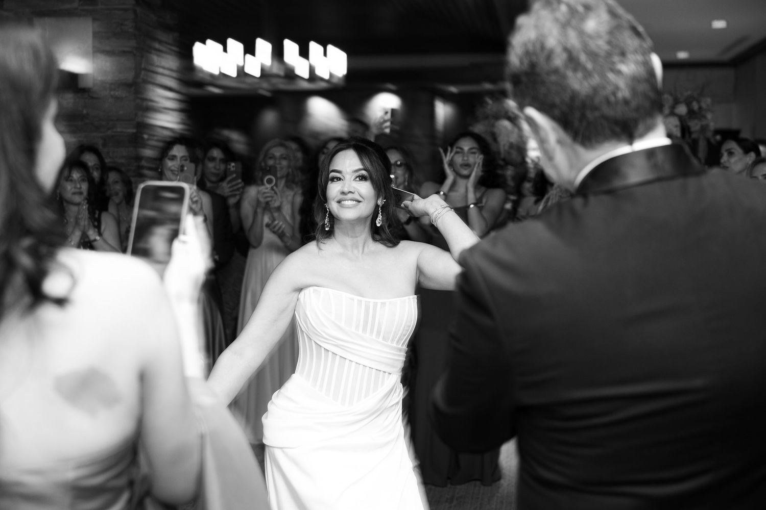 A couple dances together at their wedding reception while guests watch and take photos in black and white.