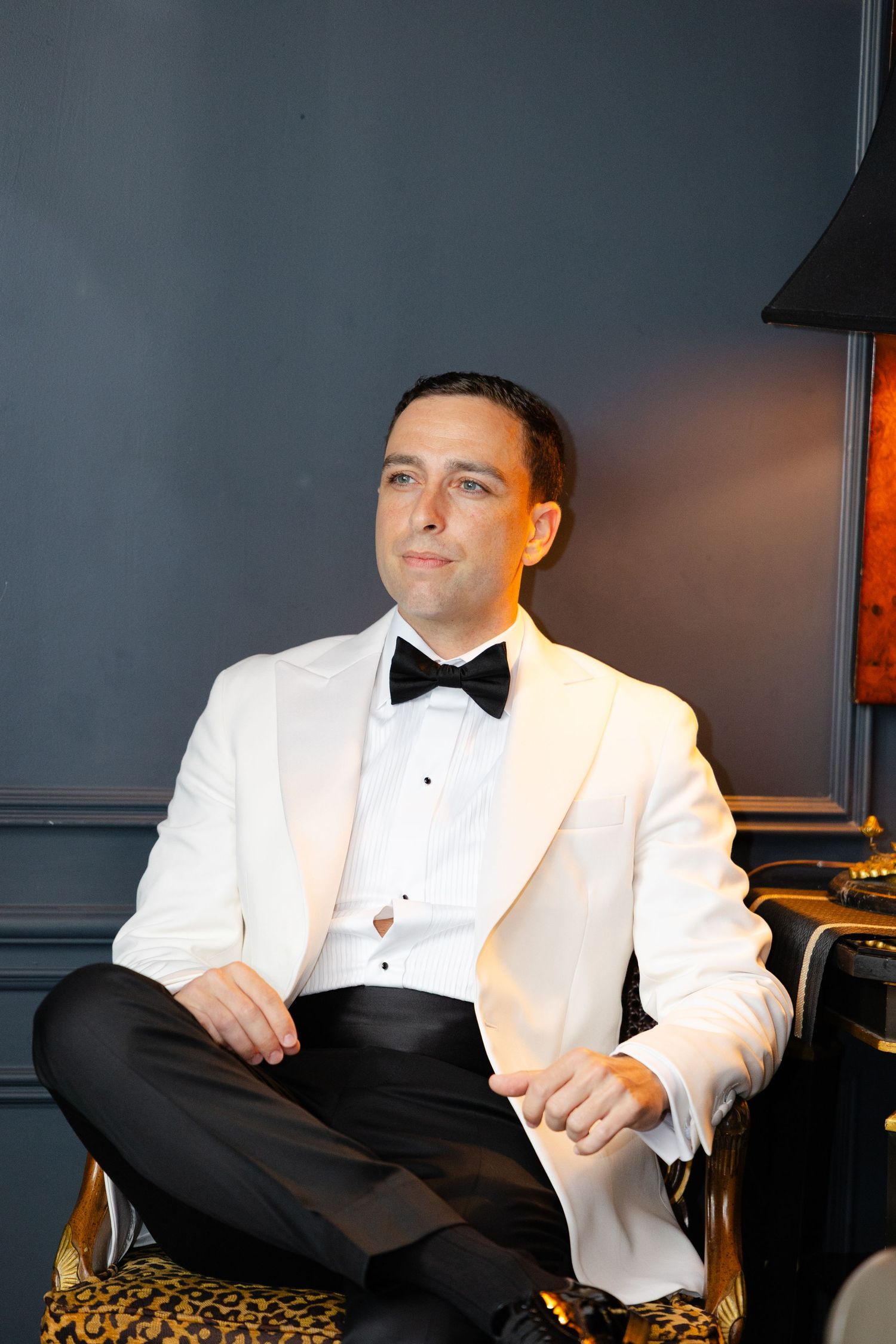A person in a white tuxedo jacket sits in an elegant chair against a dark background in a series of poses.
