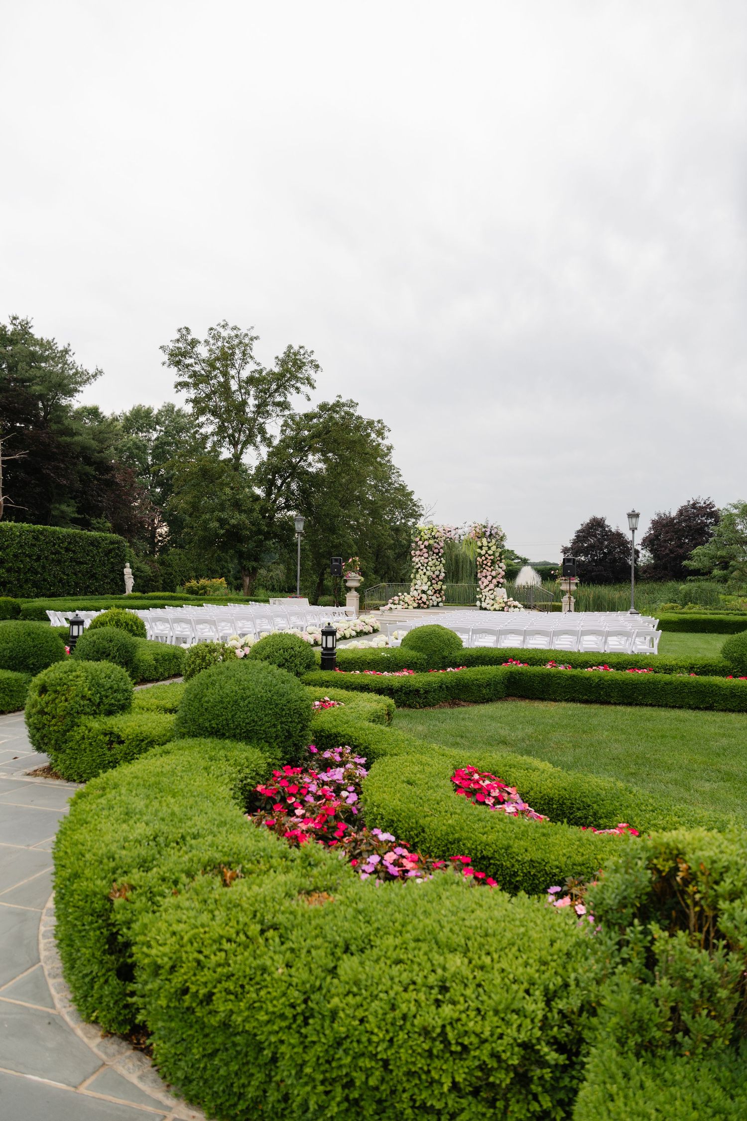 A series of manicured green hedges and vibrant red flowers line a winding garden path under an overcast sky.